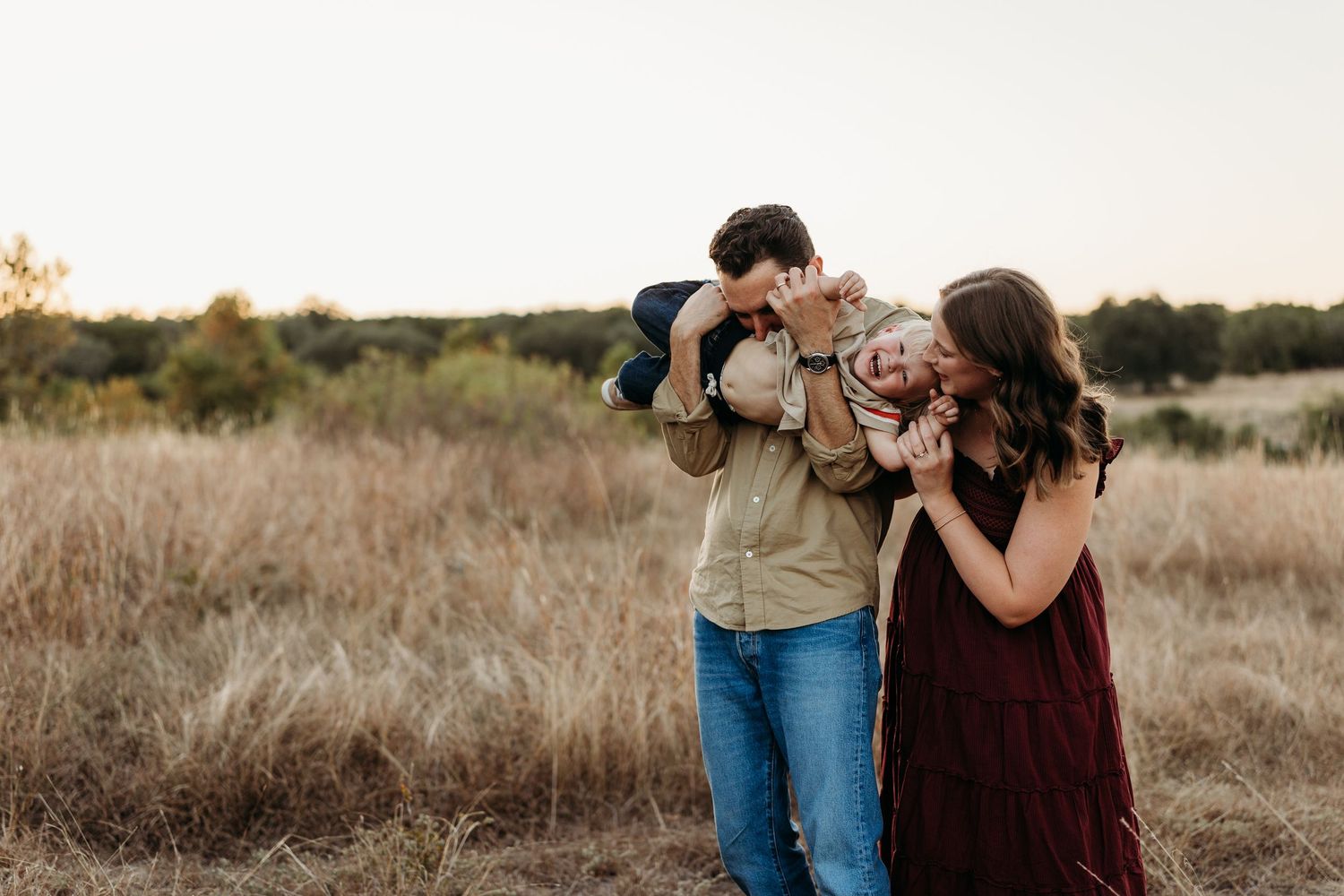 Parents playfully lifting and kissing their toddler in a warm golden field at sunset.