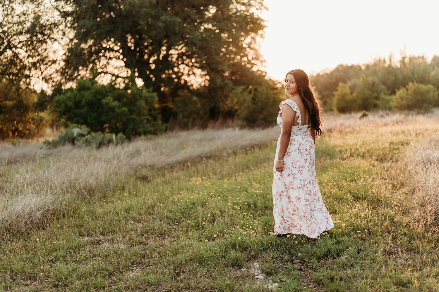 Woman in floral maxi dress standing in golden sunlit meadow surrounded by lush green trees.