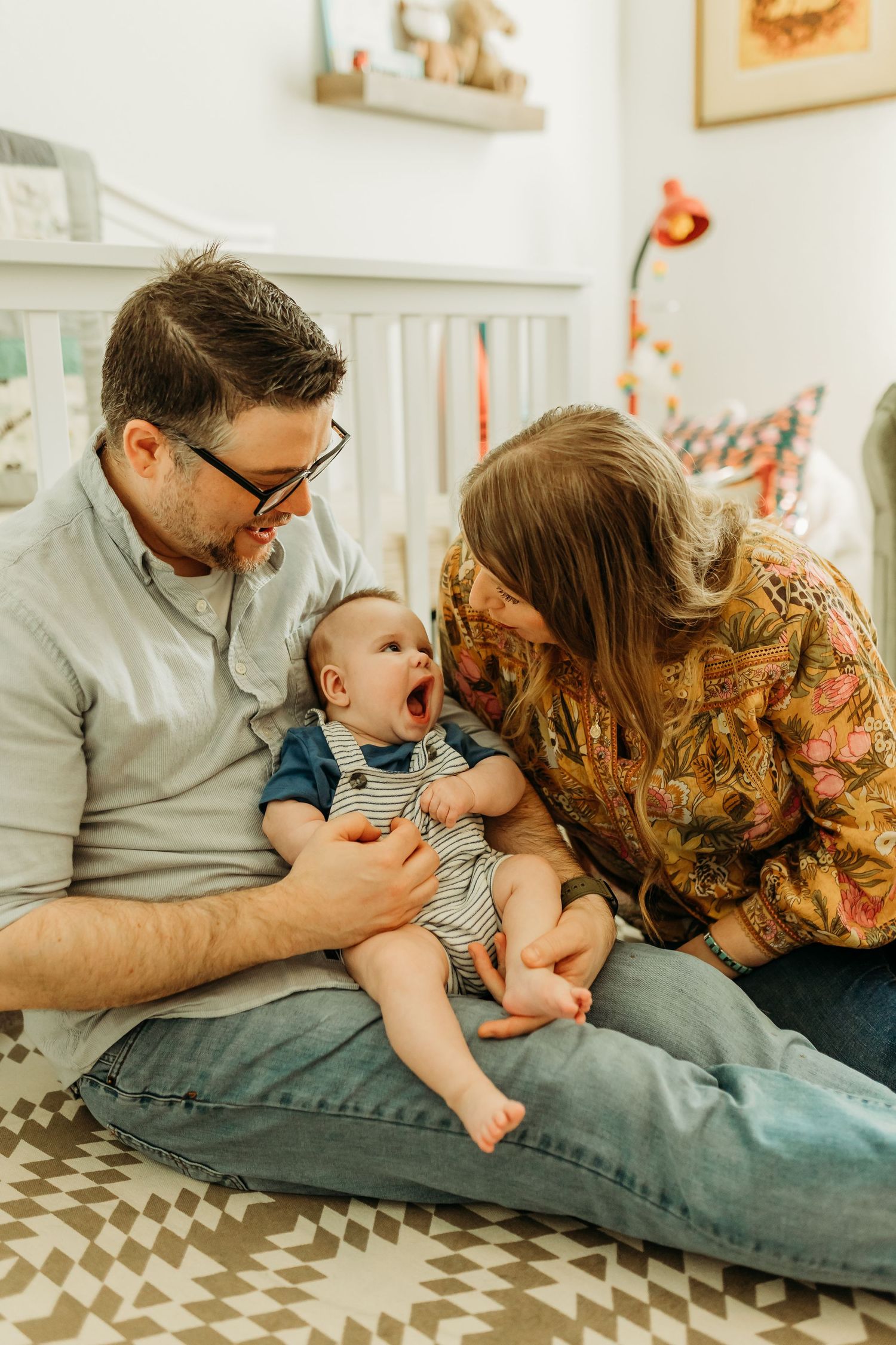 Parents sitting on nursery floor smiling at yawning baby between them.