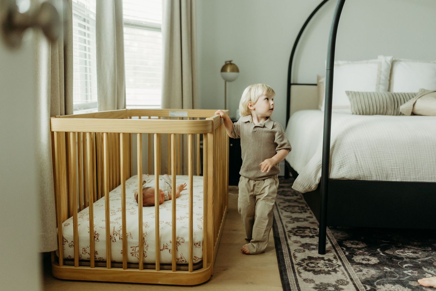 Curious toddler standing beside a wooden baby crib with sleeping newborn in a softly decorated nursery.