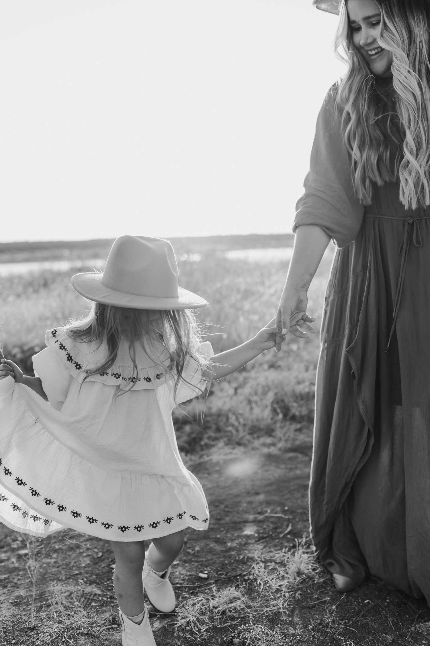 Black and white photo of a mother and young daughter in boho dresses and hats holding hands in a field.