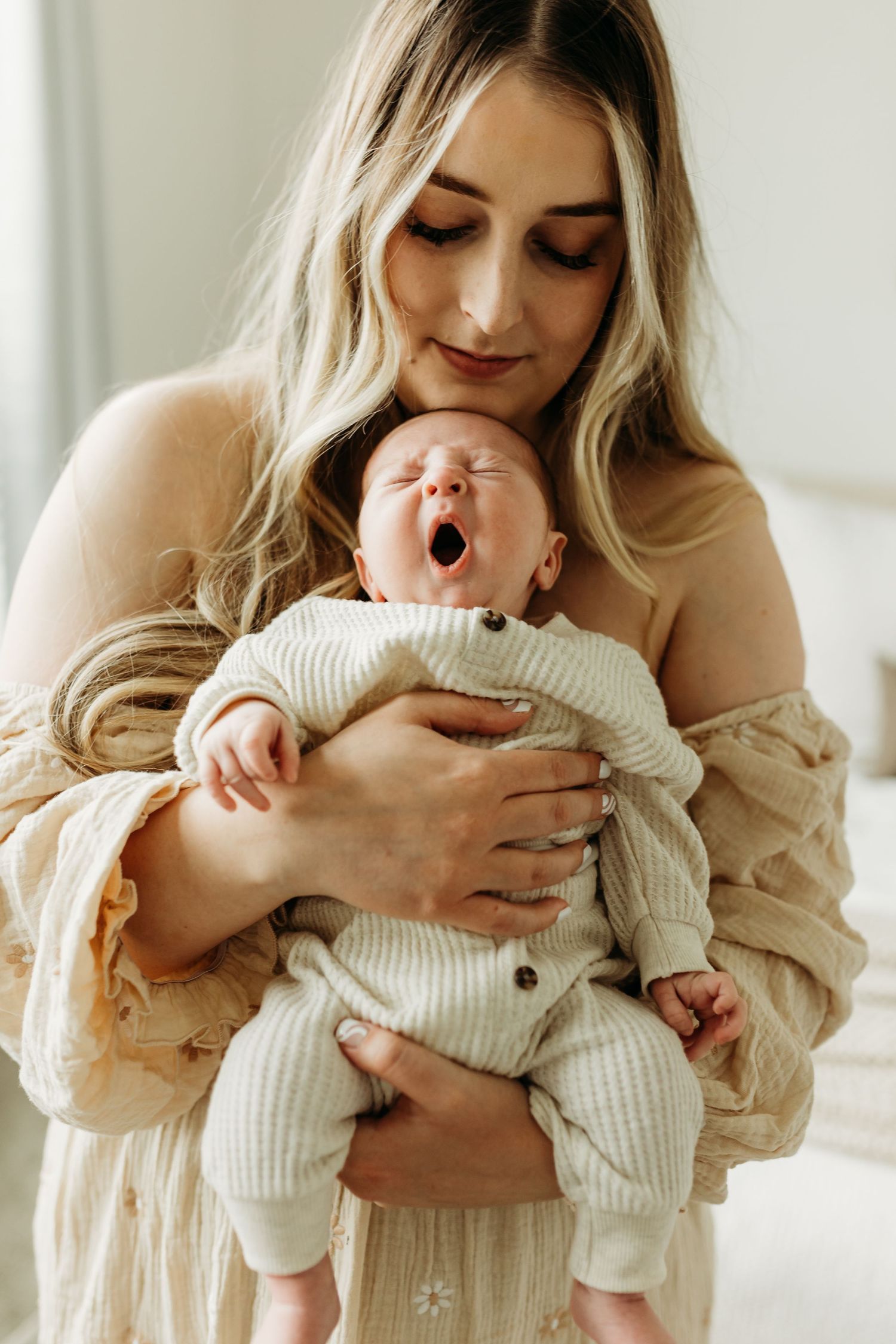 Mother tenderly holds yawning newborn baby dressed in cream ribbed outfit against her chest.