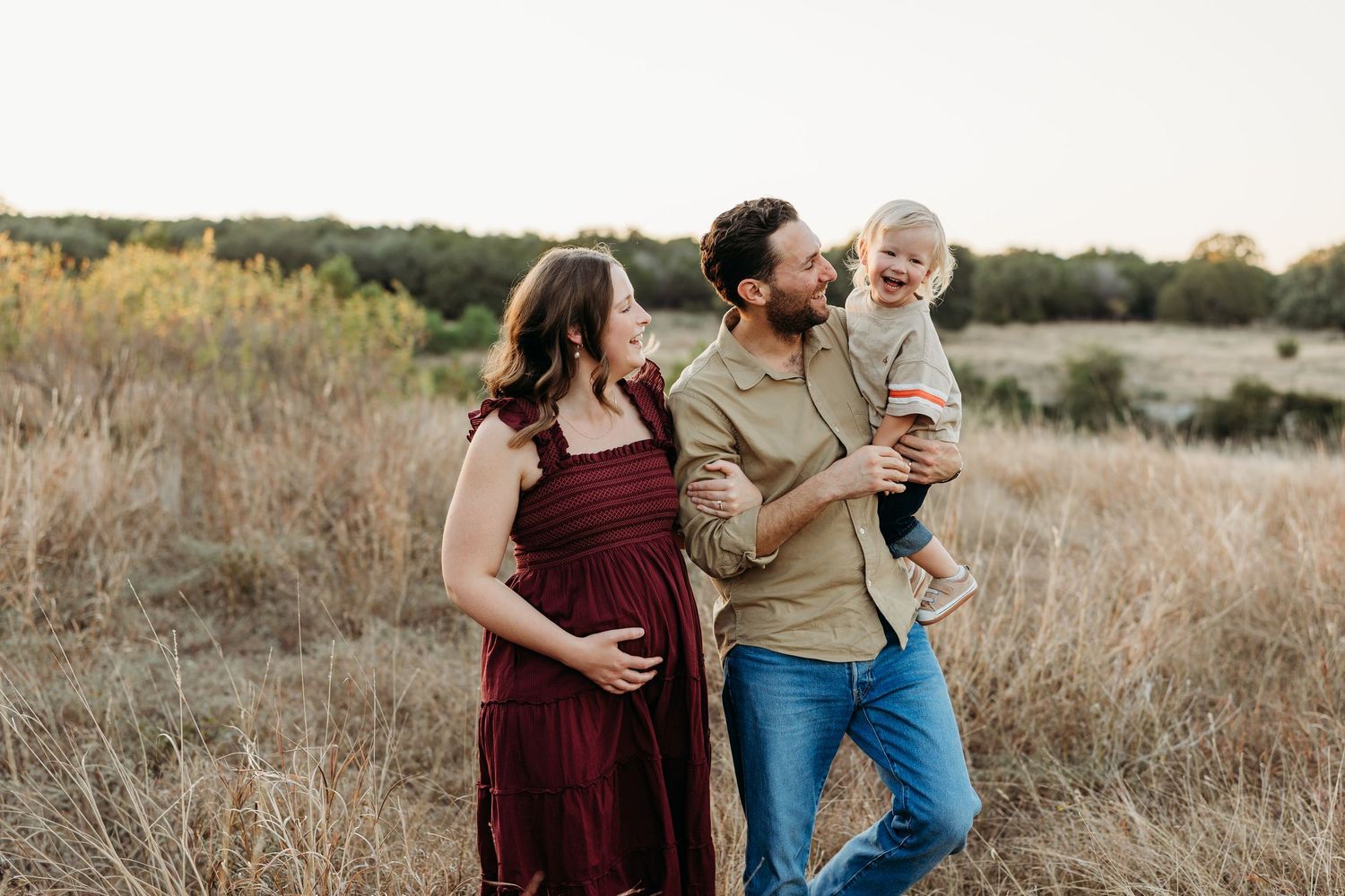 Pregnant mother and father laughing with toddler in arms through a golden sunlit field.