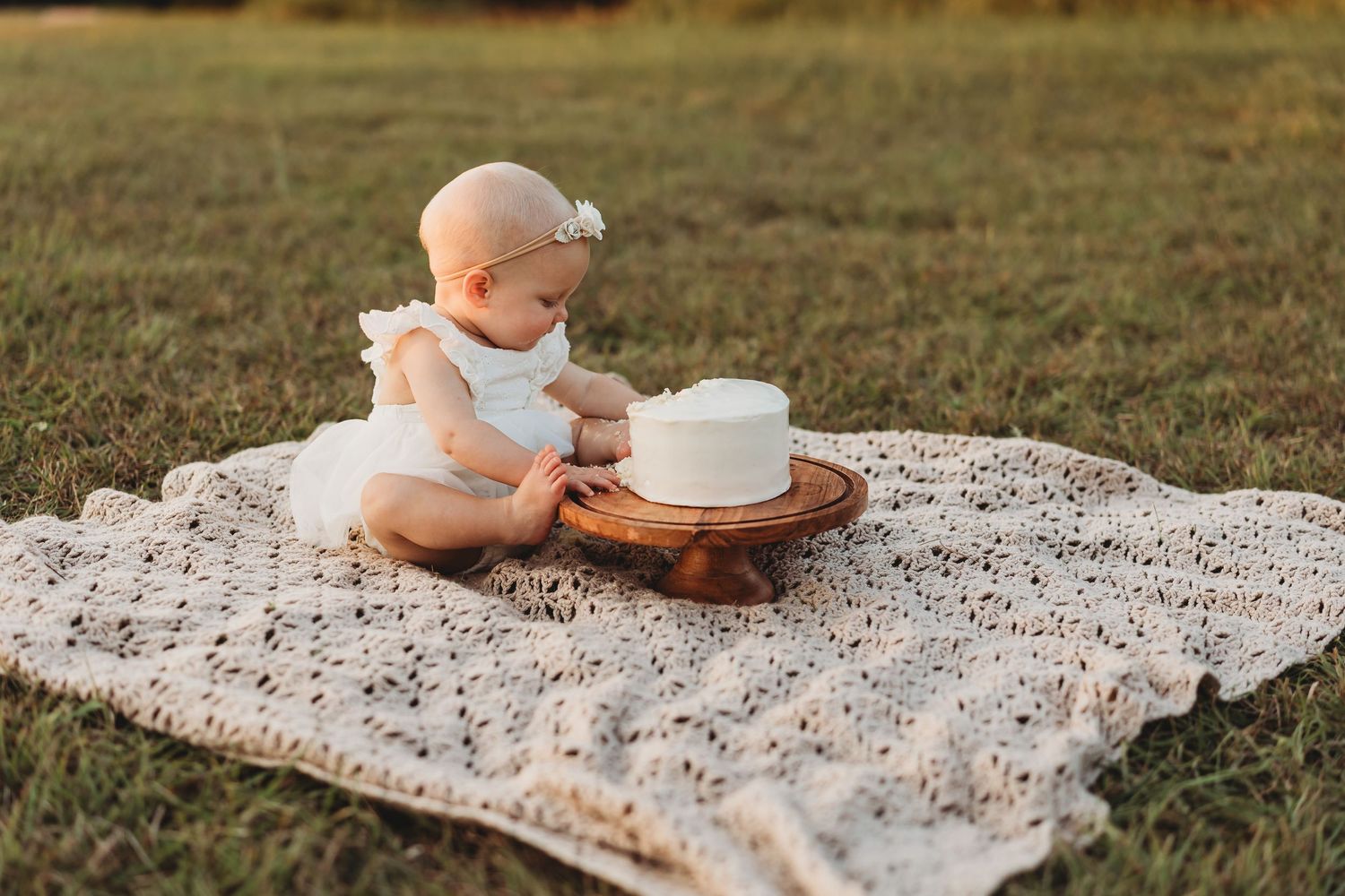Baby girl in white dress sitting on crochet blanket with first birthday smash cake.