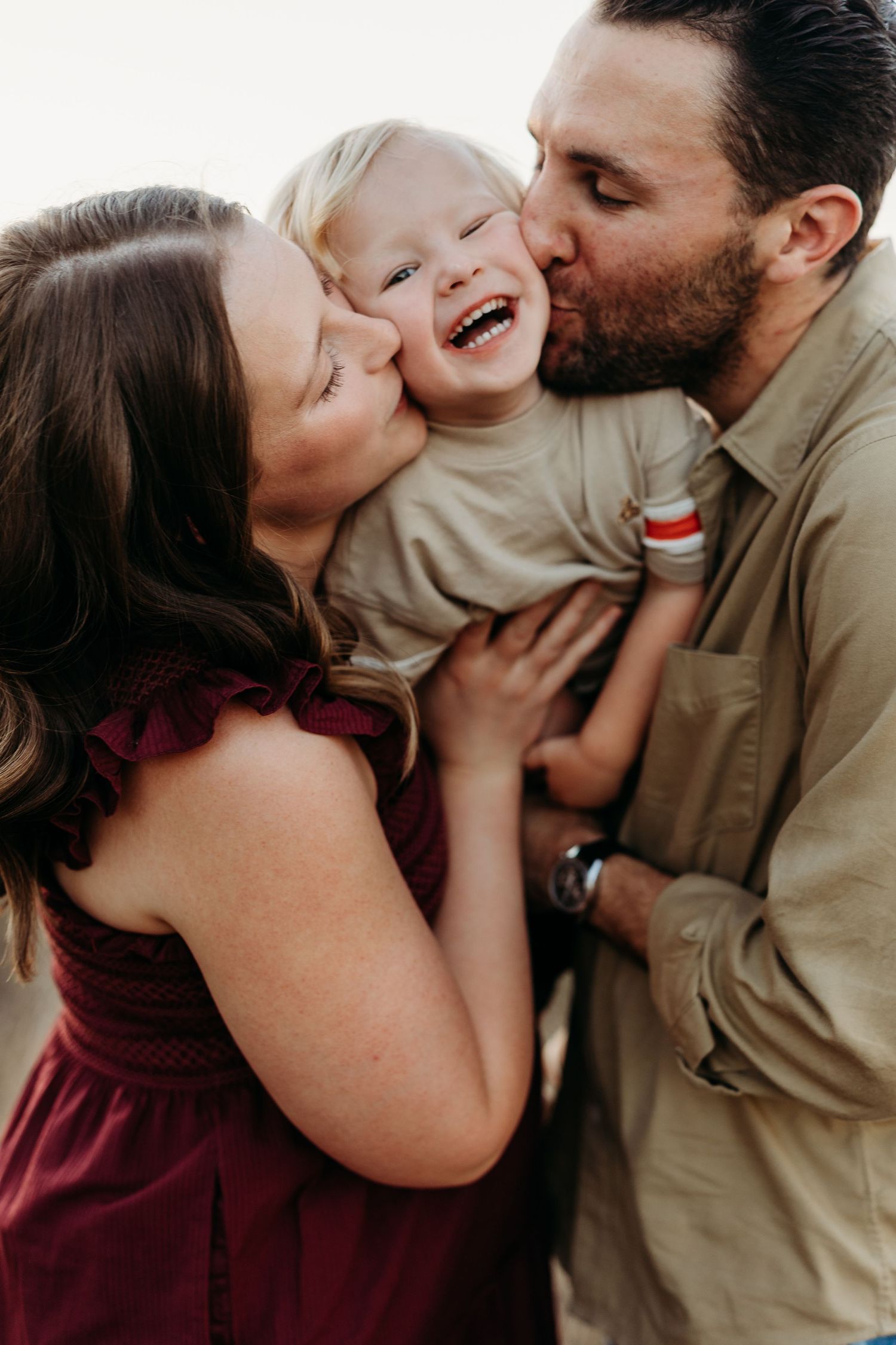 Parents lovingly kissing their giggling toddler on both cheeks during an outdoor family portrait.