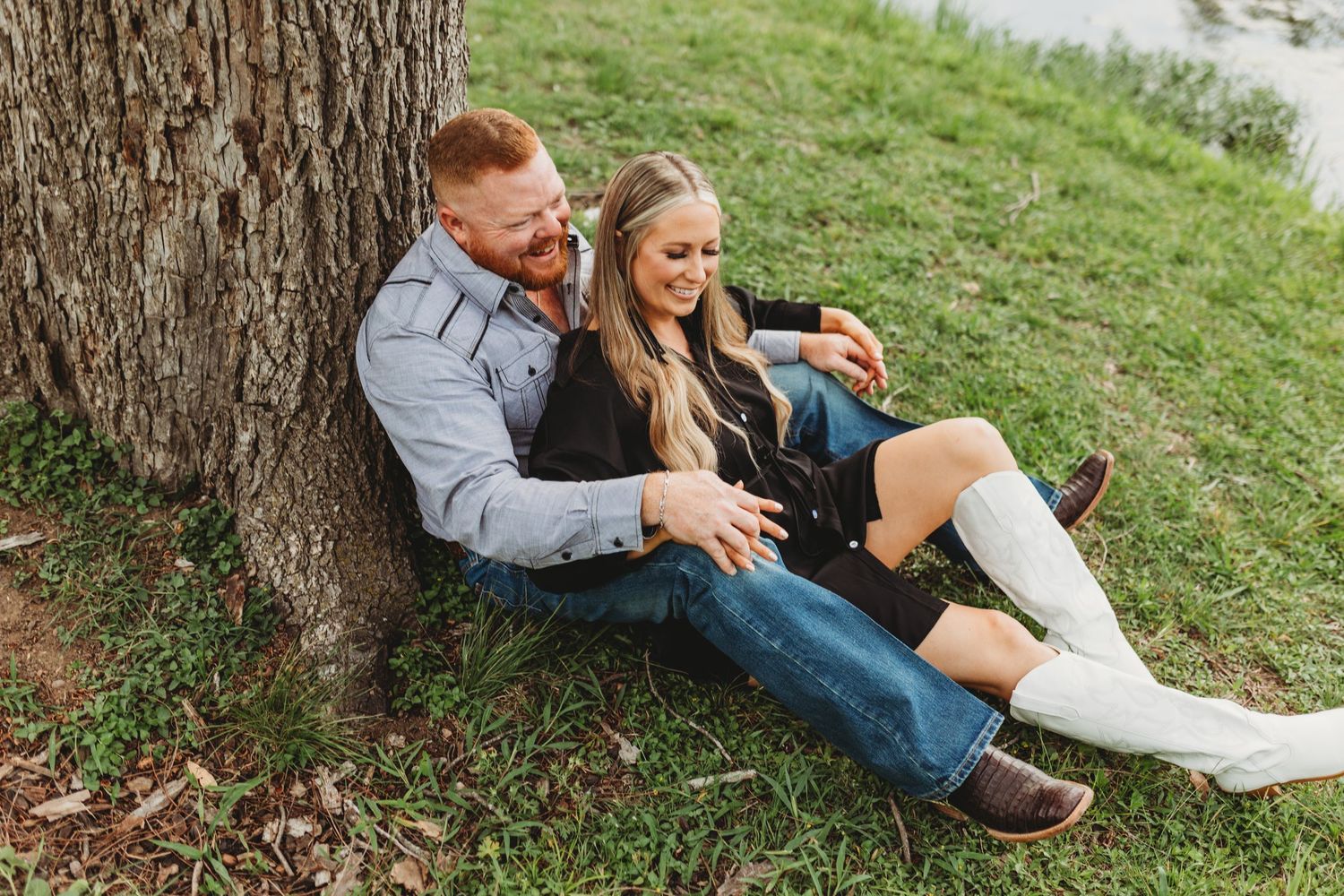 A smiling couple relaxes against a tree on green grass, sharing a joyful moment outdoors.