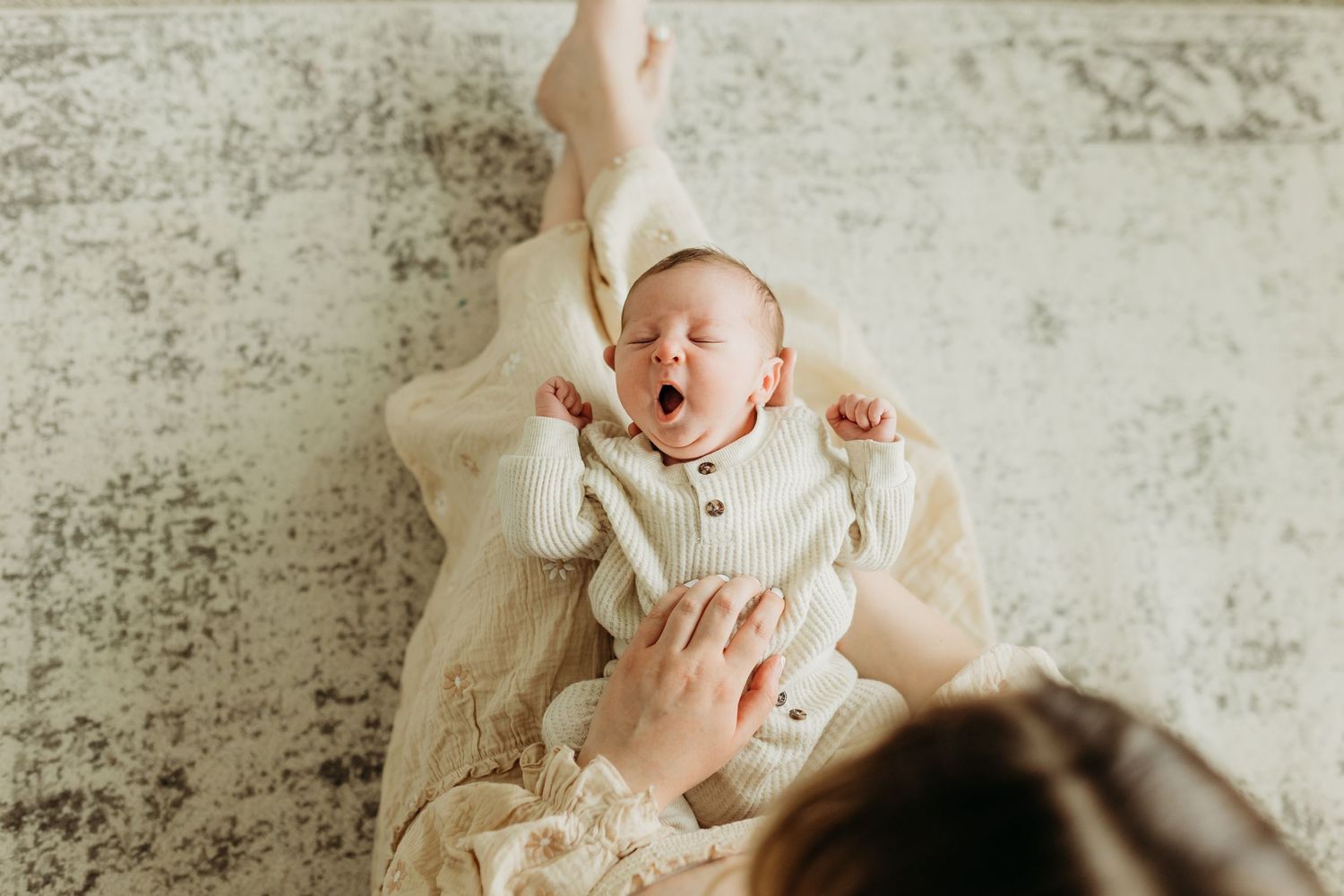 Mother watches newborn baby yawn while lying on a cream blanket on the floor.