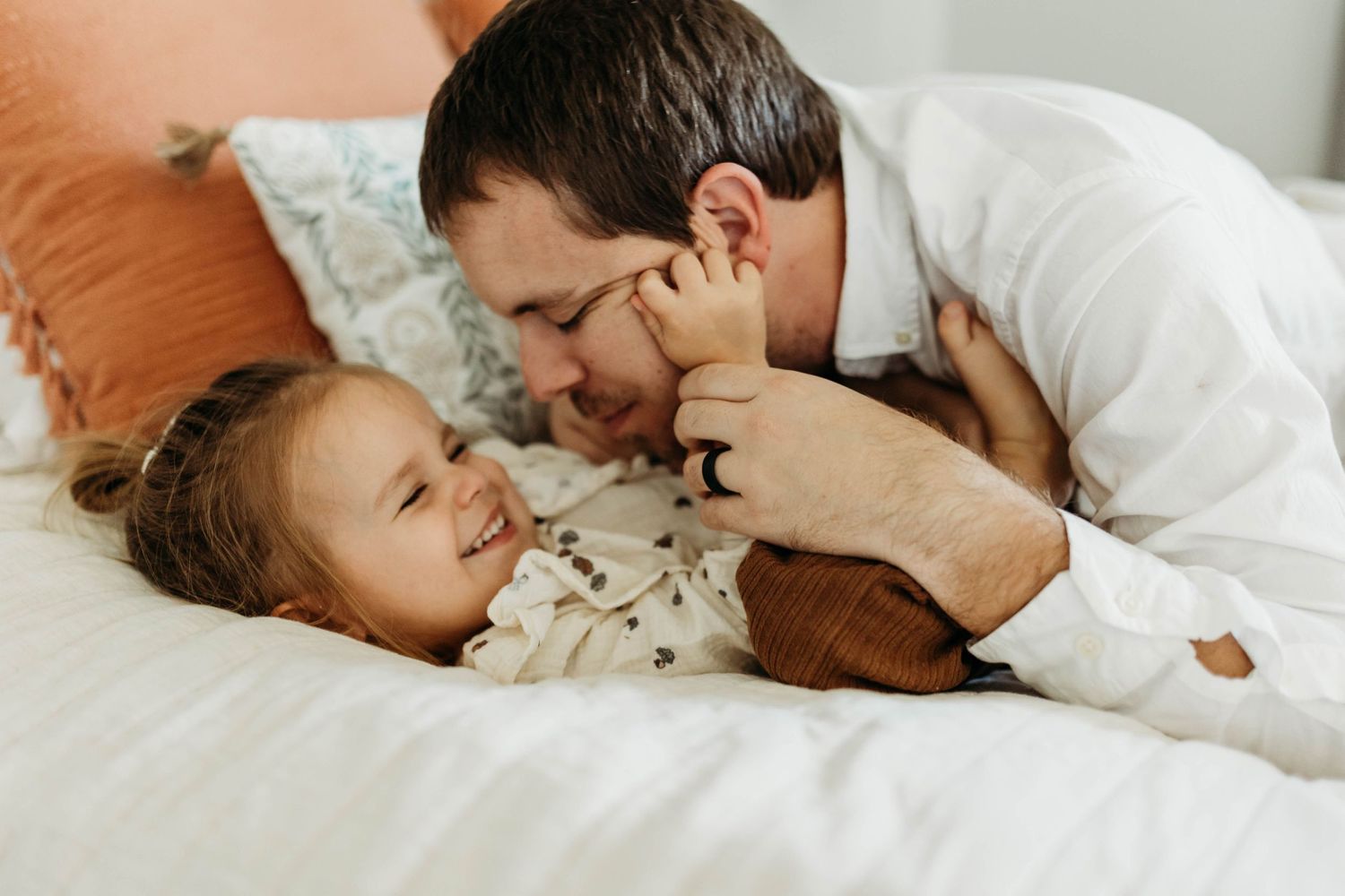 Father playfully nuzzling smiling toddler daughter while lying together on white bedding.