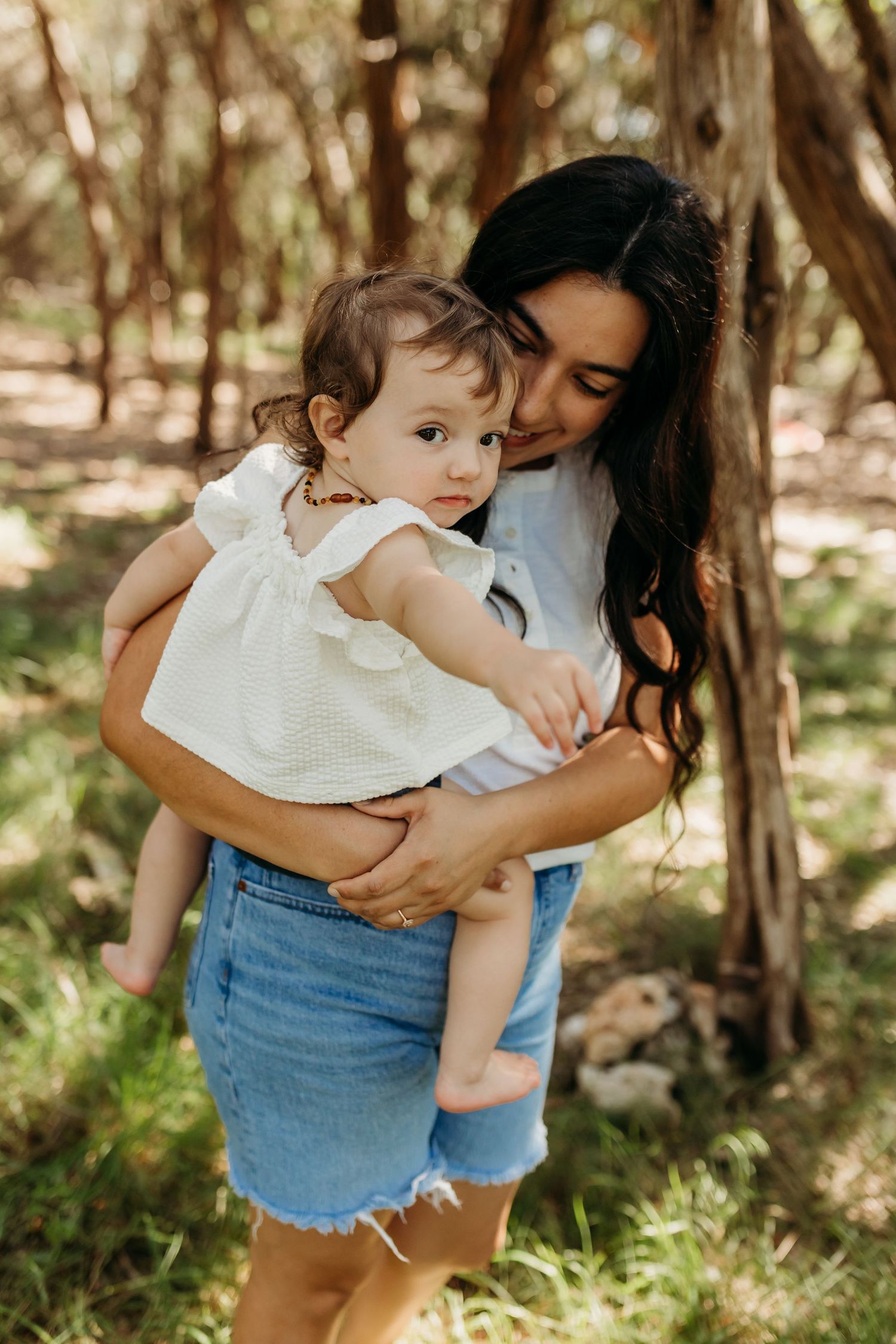 A dark-haired mother tenderly holds her baby girl against a tree in a sunlit forest setting.