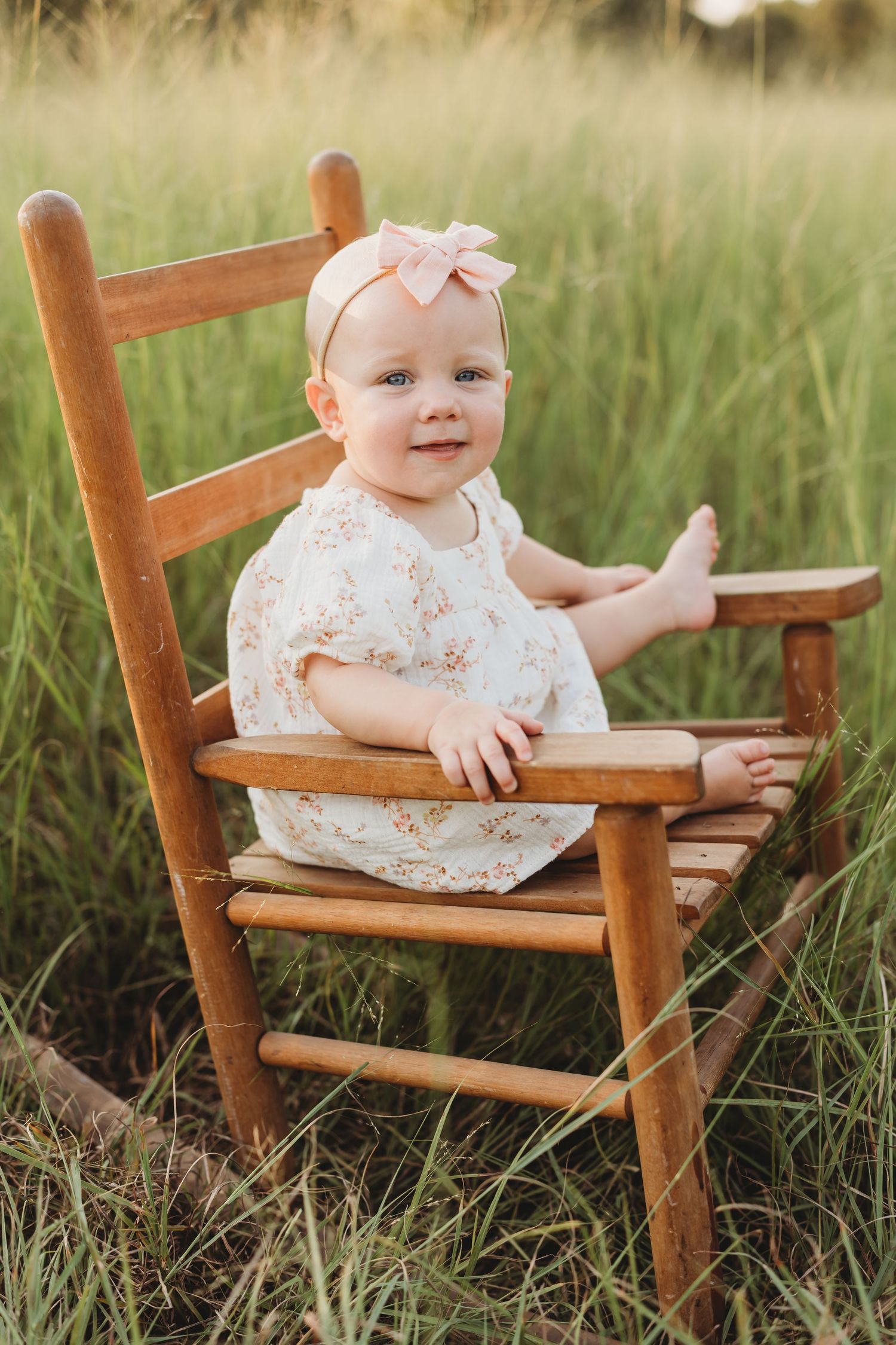 Happy baby girl in floral dress sitting on small wooden chair in sunny green field.