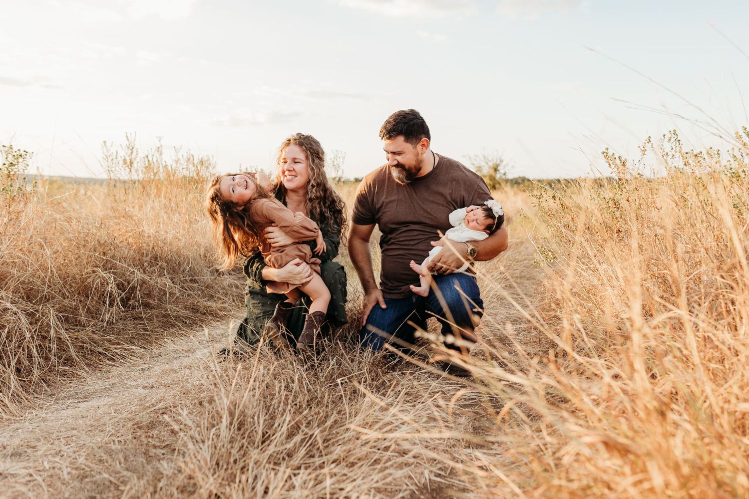 Happy family of four laughing together on a path through golden grass fields at sunset.