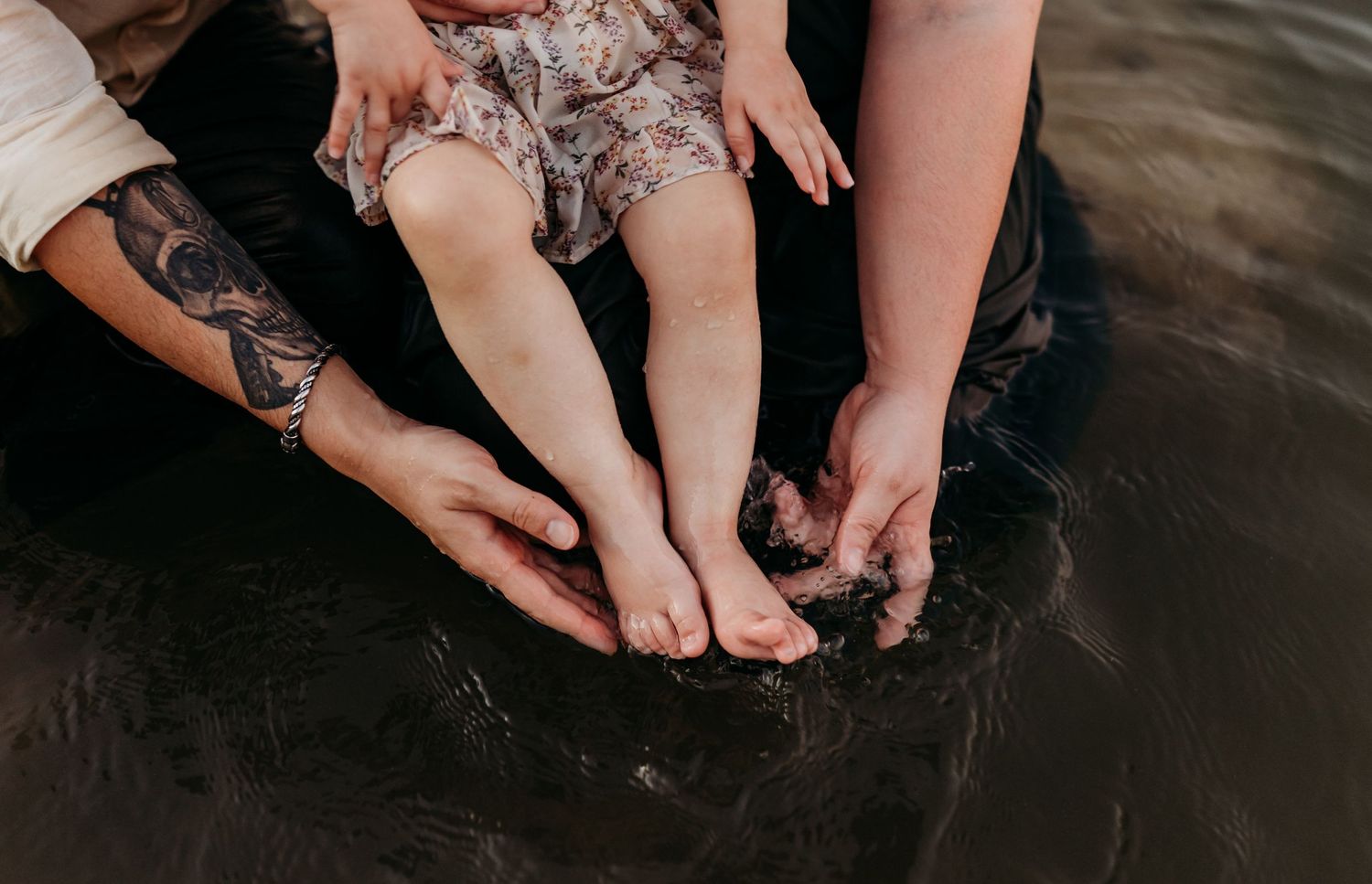 Parents with tattoos dipping baby's feet into dark water during outdoor family session.