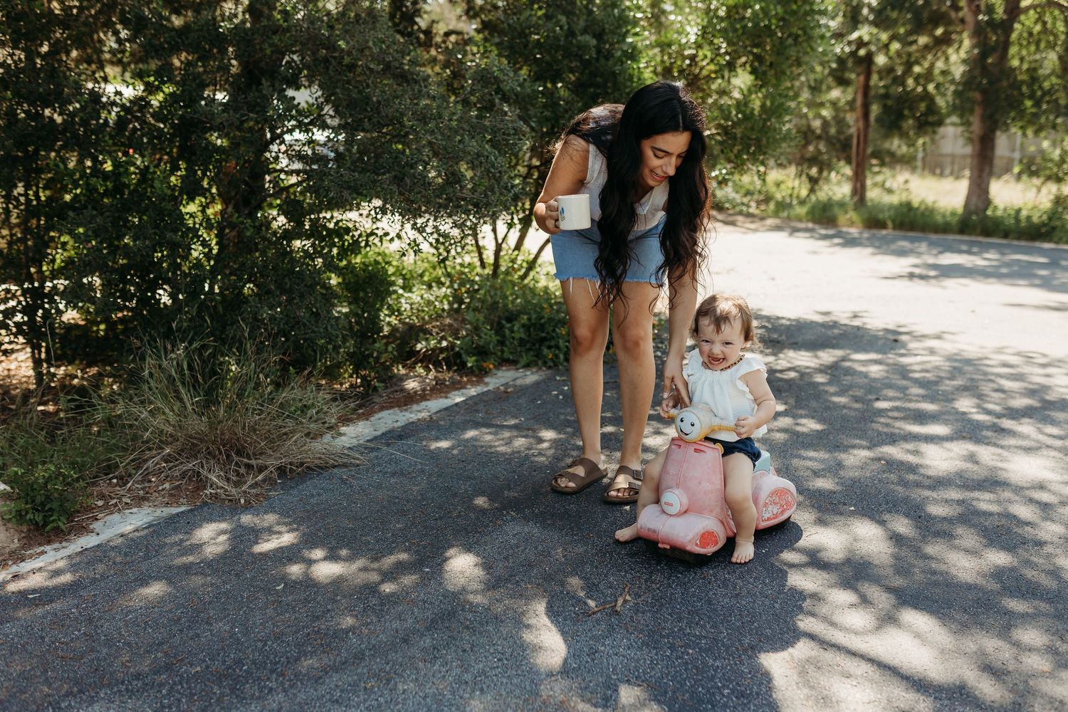 A dark-haired mother watches her toddler ride a pink toy scooter on a tree-lined pathway.