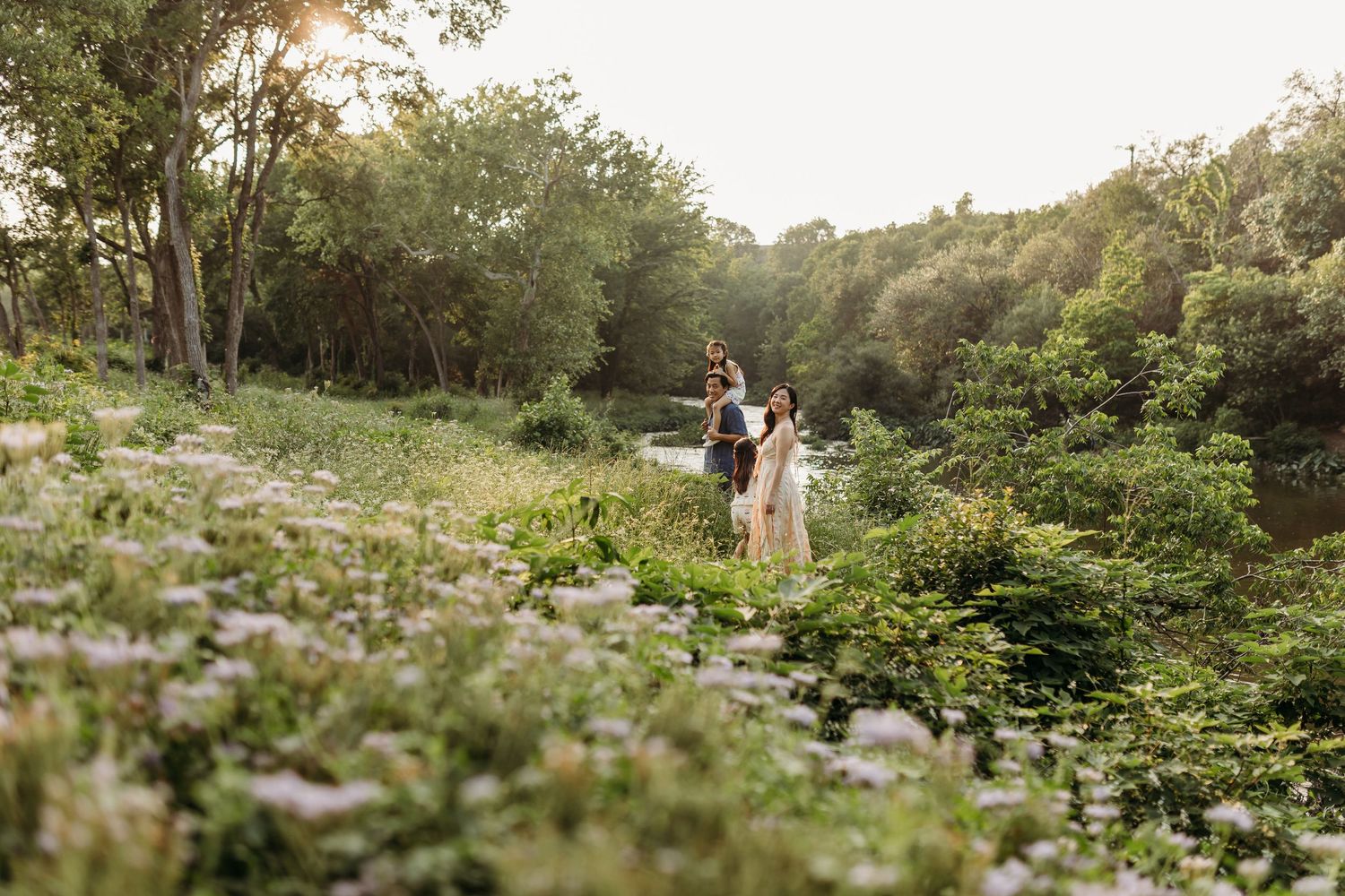 Family standing together among wildflowers by a sunlit creek in a lush green outdoor setting.