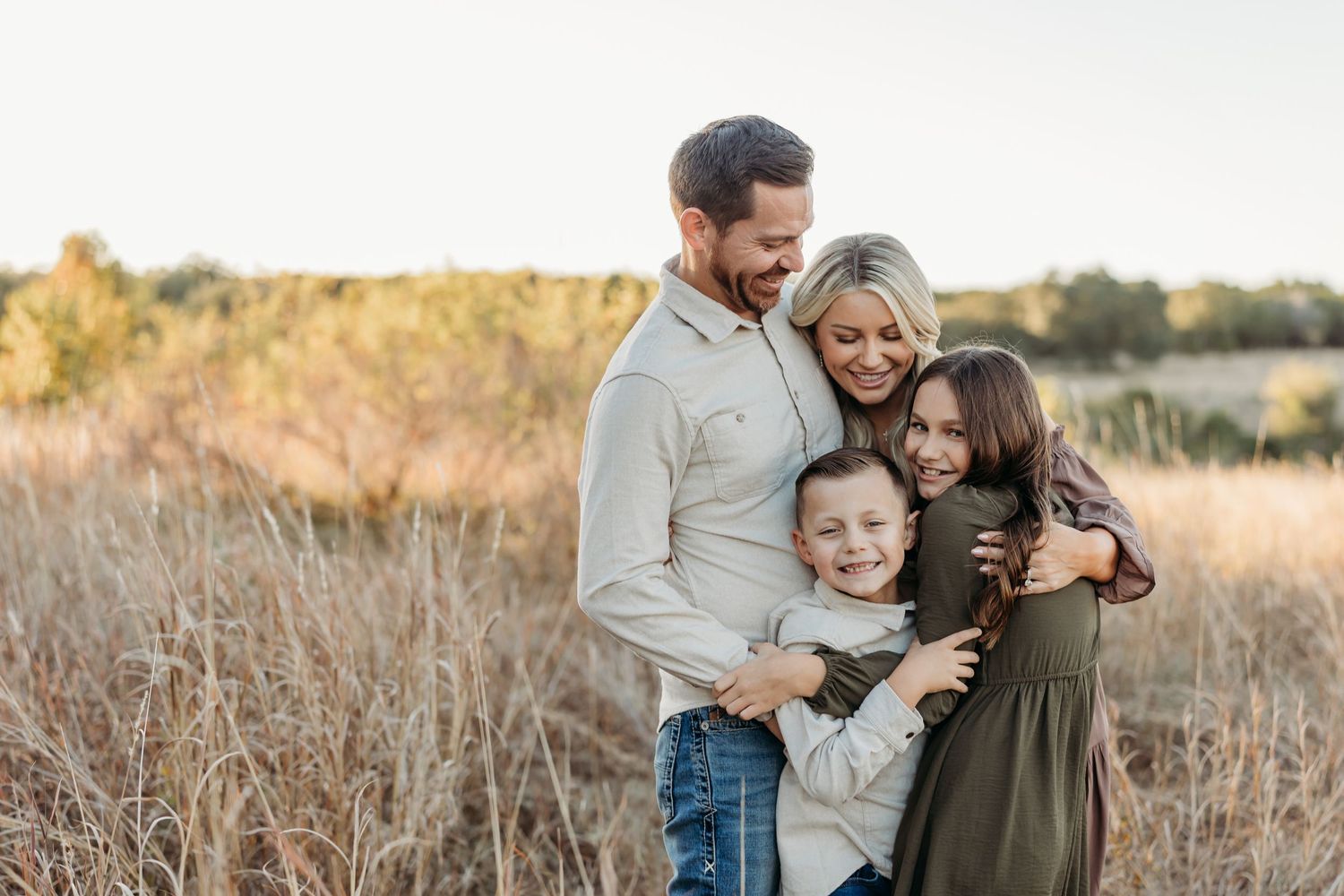 Happy family of four hugging and smiling together in a sunlit autumn field.
