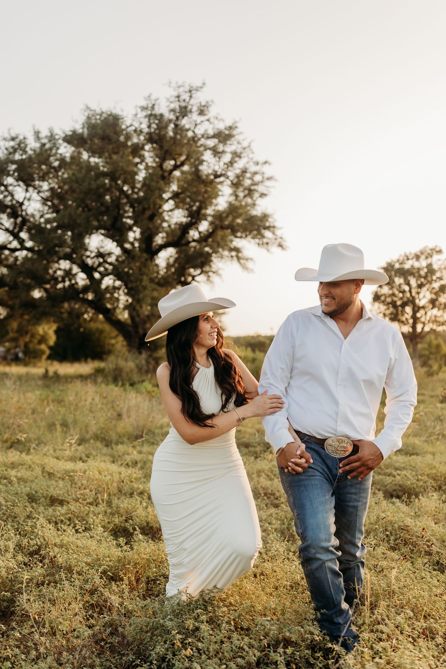 Couple in white cowboy hats walking through a sunlit Texas field.