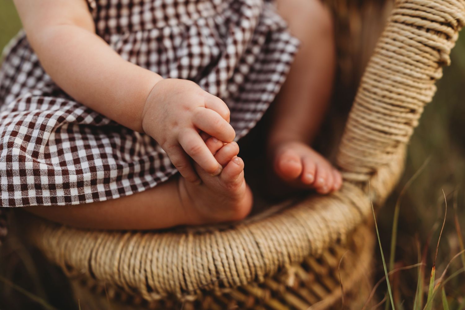 Close-up of baby hands and feet in a wicker chair, wearing a brown gingham dress.