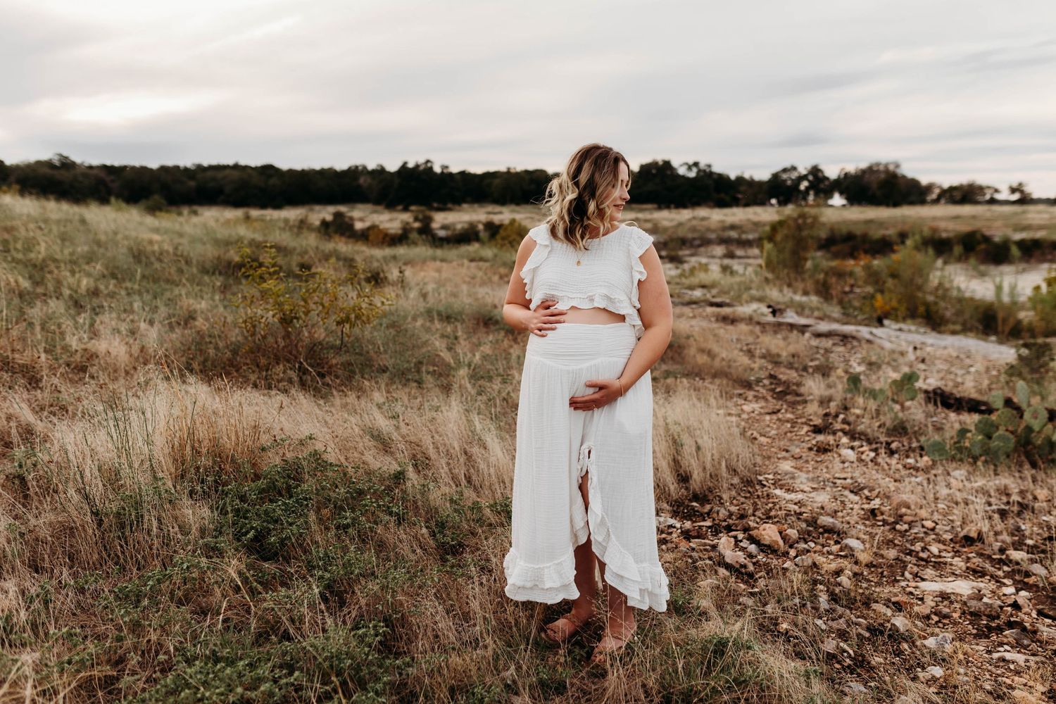 Pregnant woman in a white dress cradling her belly in an open field.