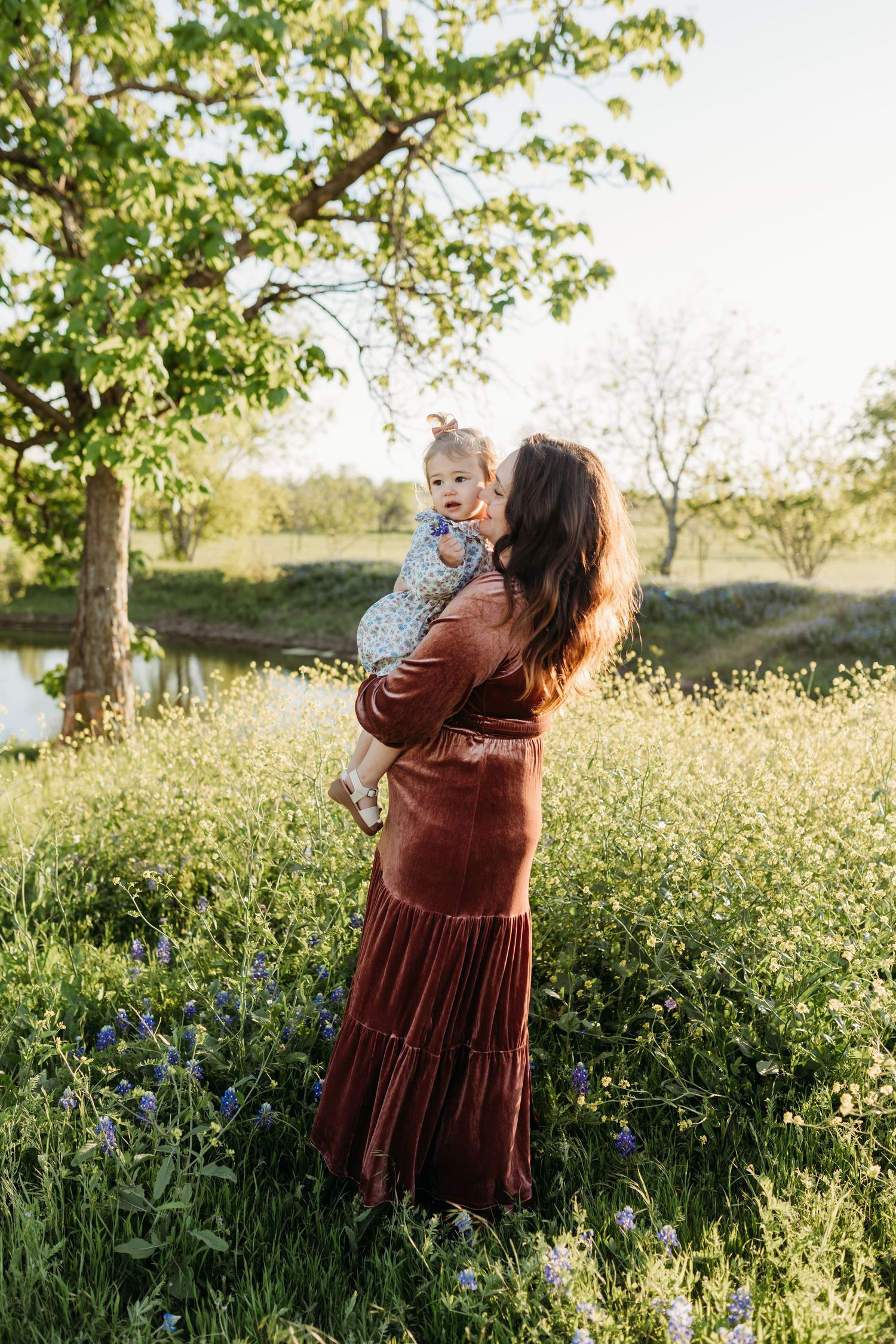 A mother in a rust velvet dress holds her baby girl in a sunlit bluebonnet field.