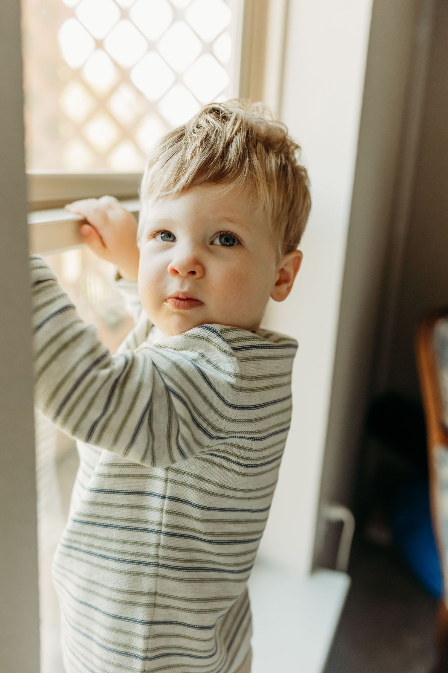 Curious blonde toddler in striped pajamas standing at a window holding the frame.