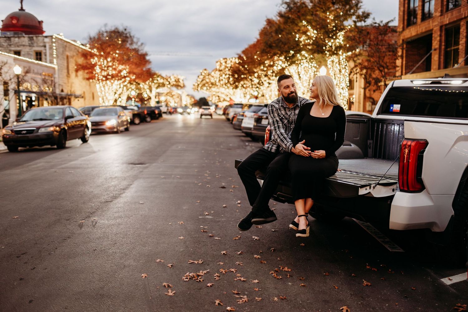 Couple sitting on truck tailgate on a festive city street with holiday lights at dusk.