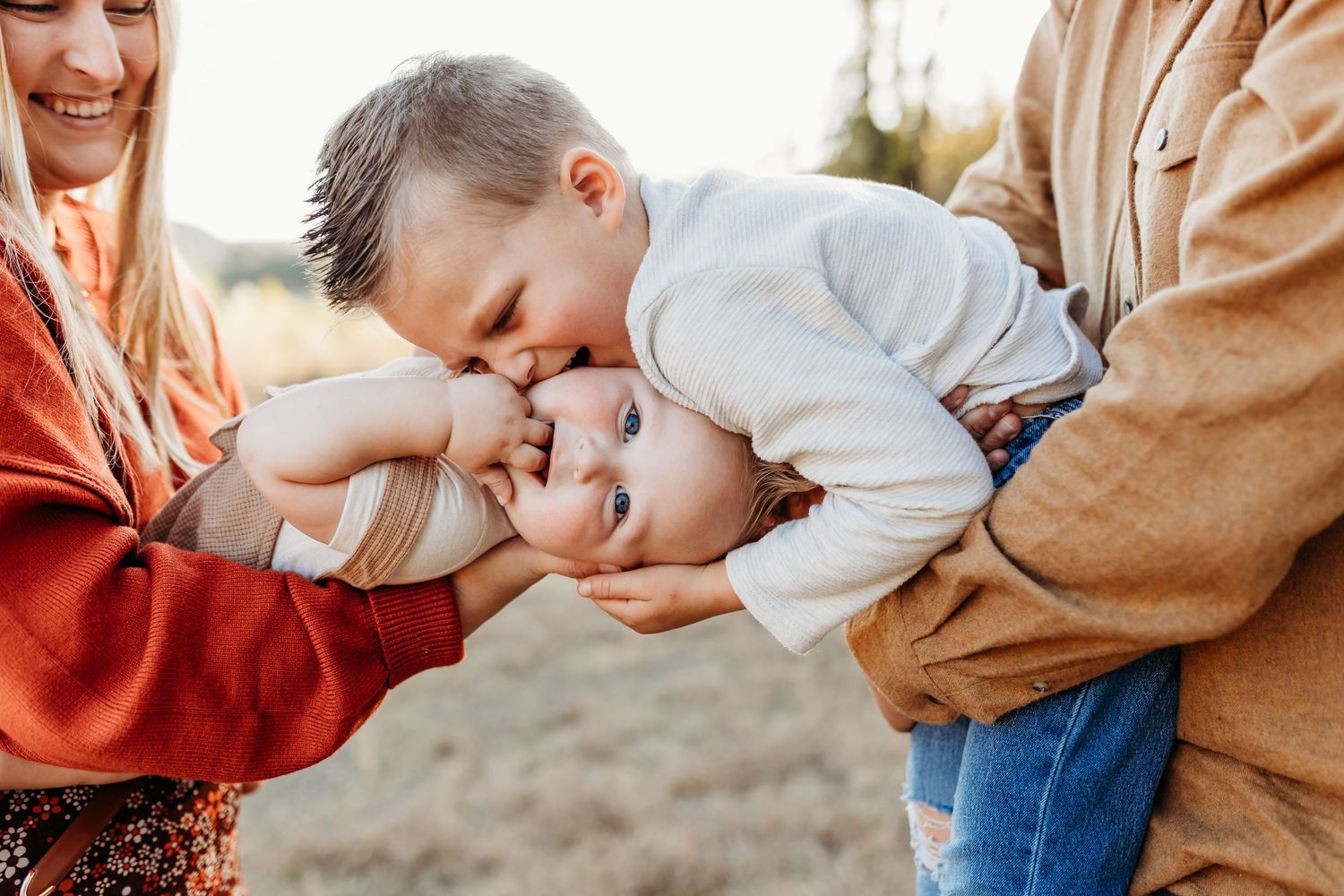 Young boy lovingly kissing smiling baby sibling held by parents outdoors.
