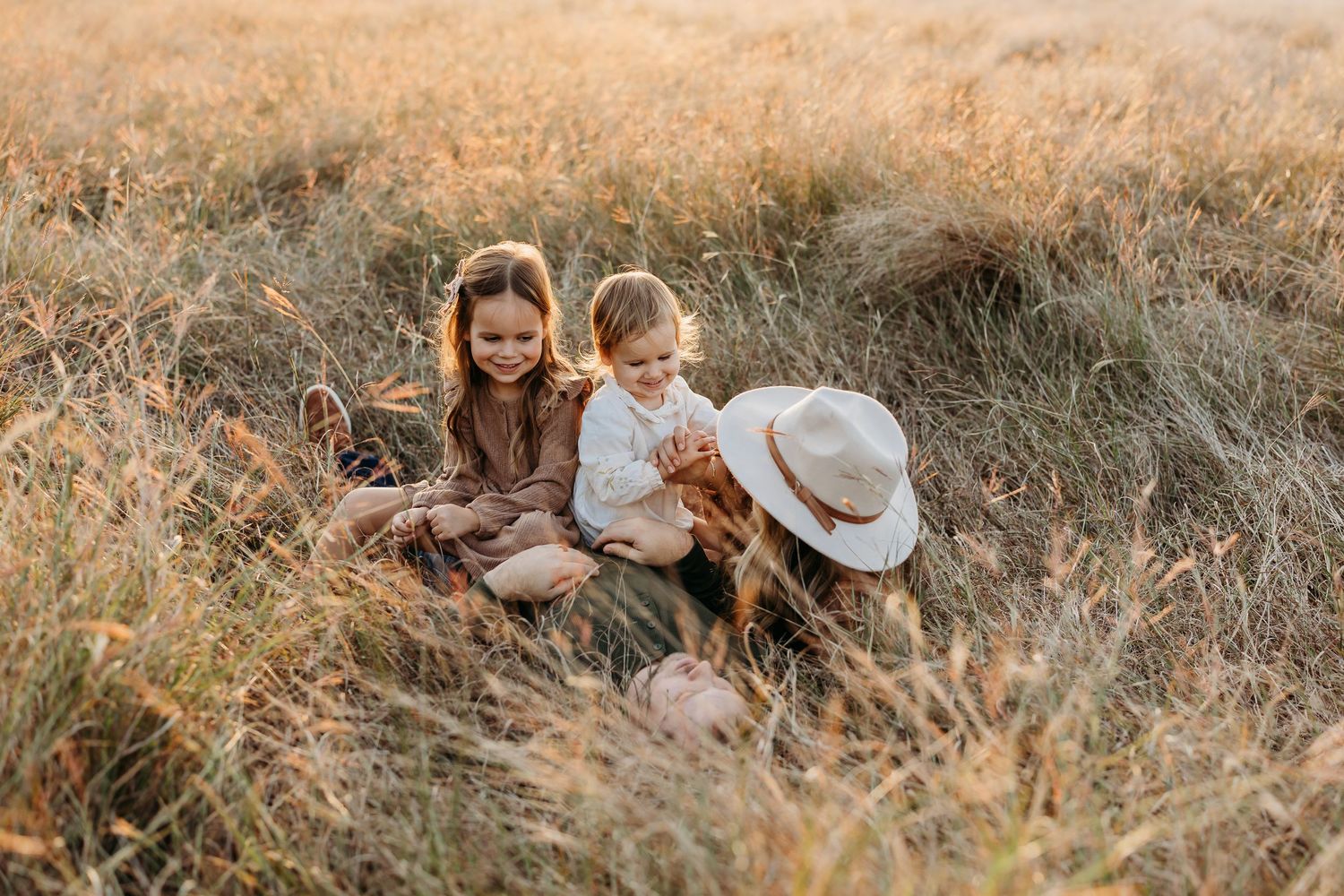 Mother sitting in golden grass field with two young daughters during warm sunset light.