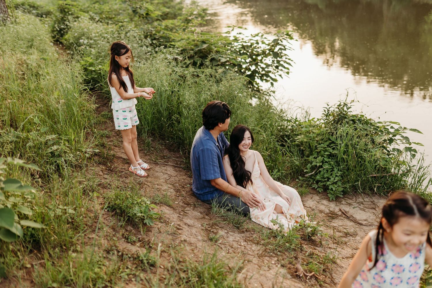 A family relaxes on a riverbank surrounded by lush greenery during a warm outdoor photo session.