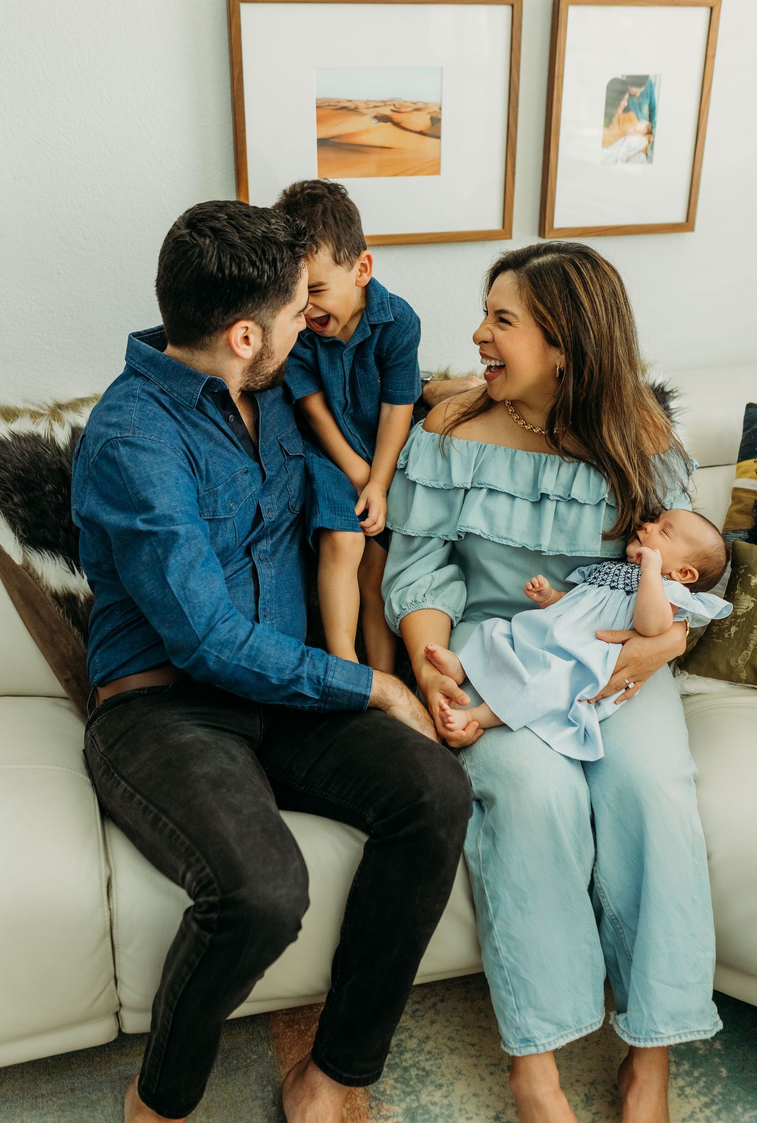 Happy family laughing together on white couch with newborn baby and young son indoors.