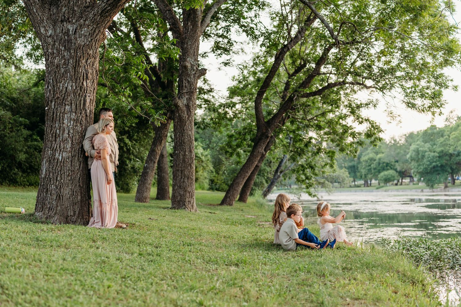 Mother in pink dress standing by large tree watching her children sit by a peaceful lakeside.