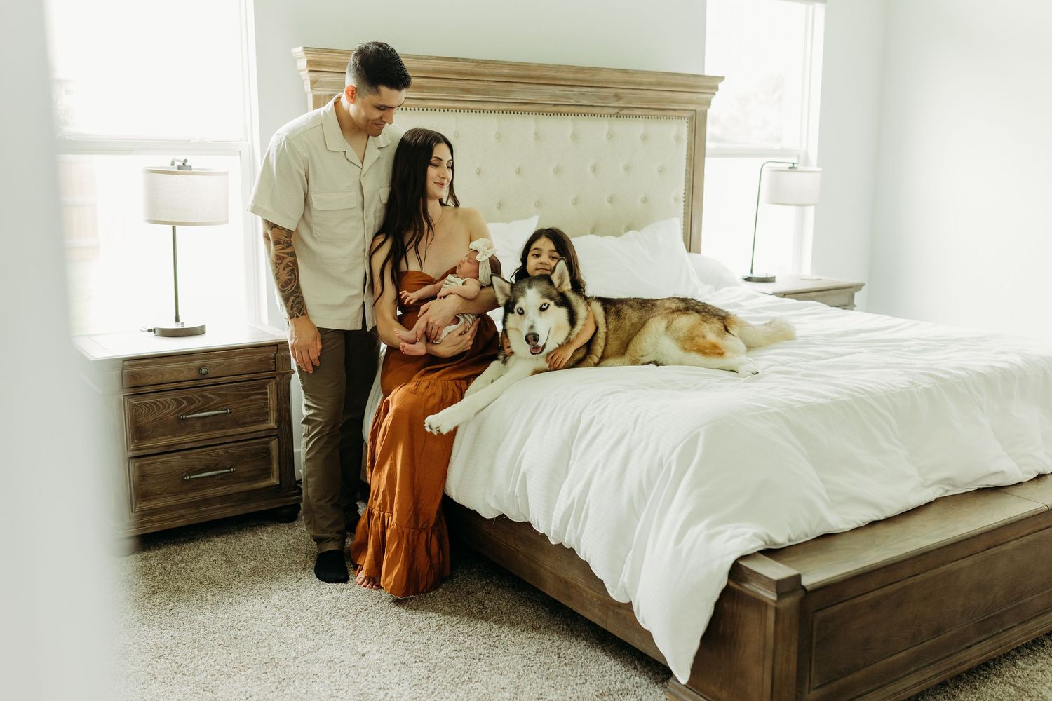 A couple and their daughter stand beside a large husky dog resting on a bed in a bright bedroom.