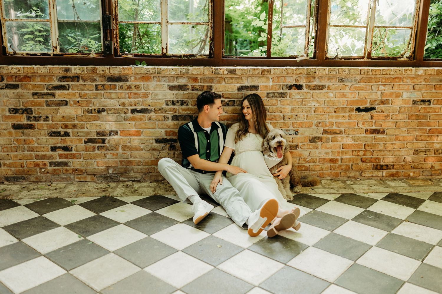 A couple sits together on a checkered floor against a brick wall, cuddling with their dog indoors.