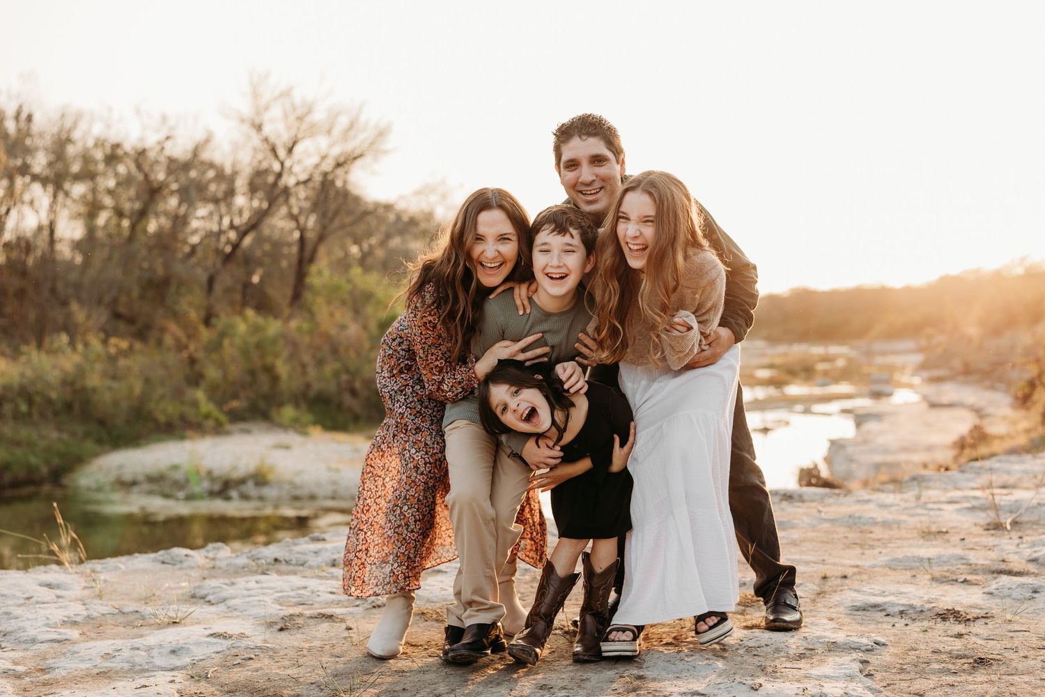 Happy family of five hugging and smiling together on rocky outdoor terrain at golden hour.