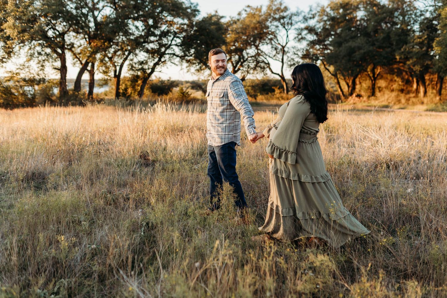 A couple holds hands and walks through a golden sunlit grassy field.