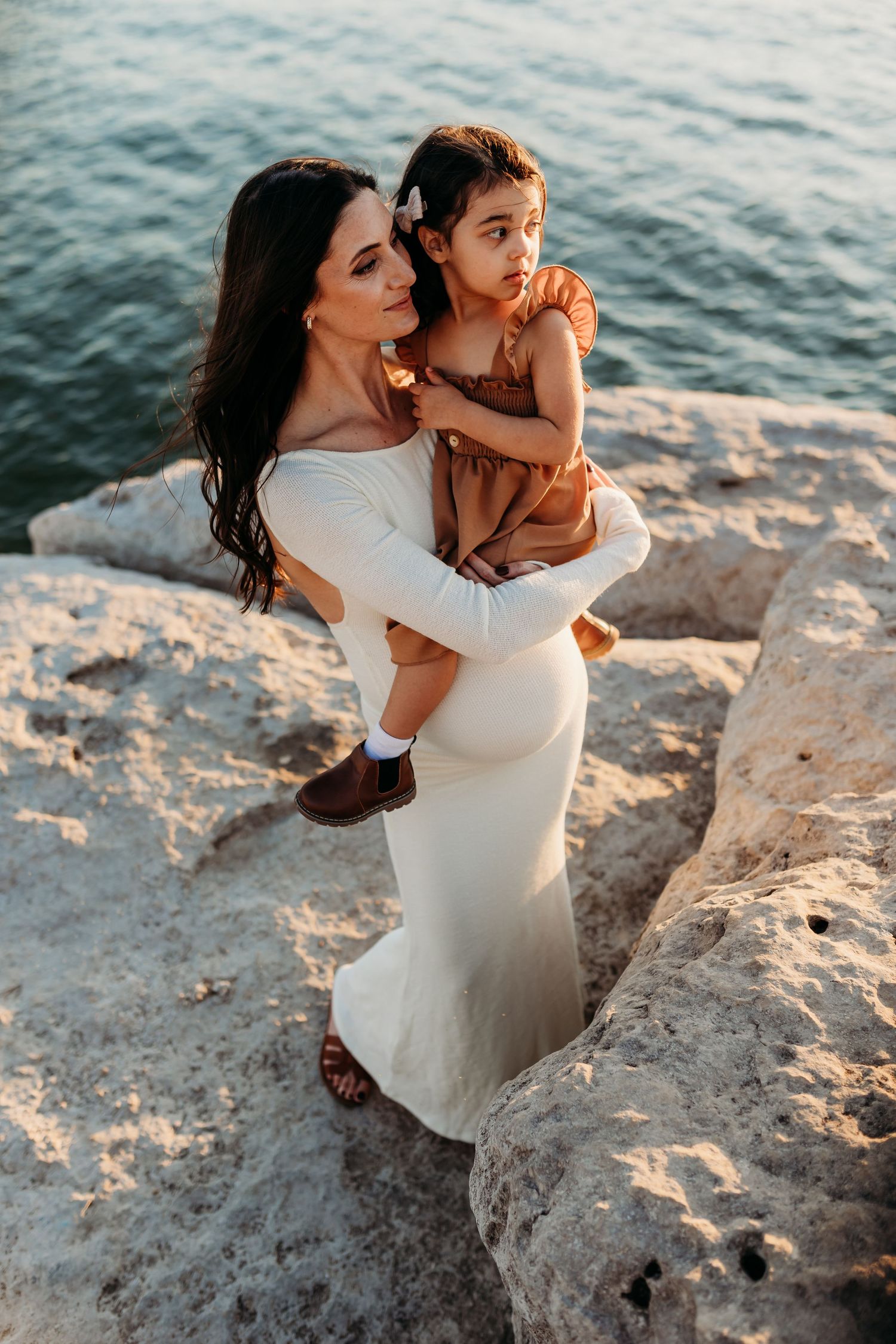 A mother in a white dress holds her young daughter on rocky cliffs by the ocean at golden hour.