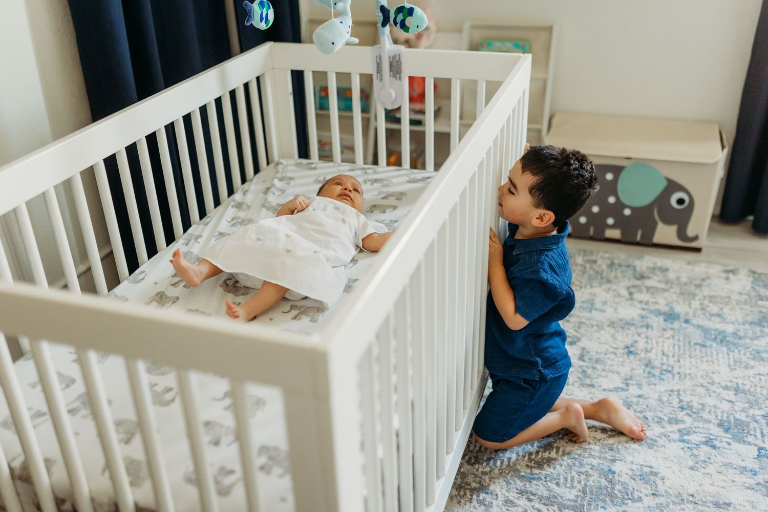 Young boy kneeling beside white crib watching over sleeping newborn sibling in nursery room.