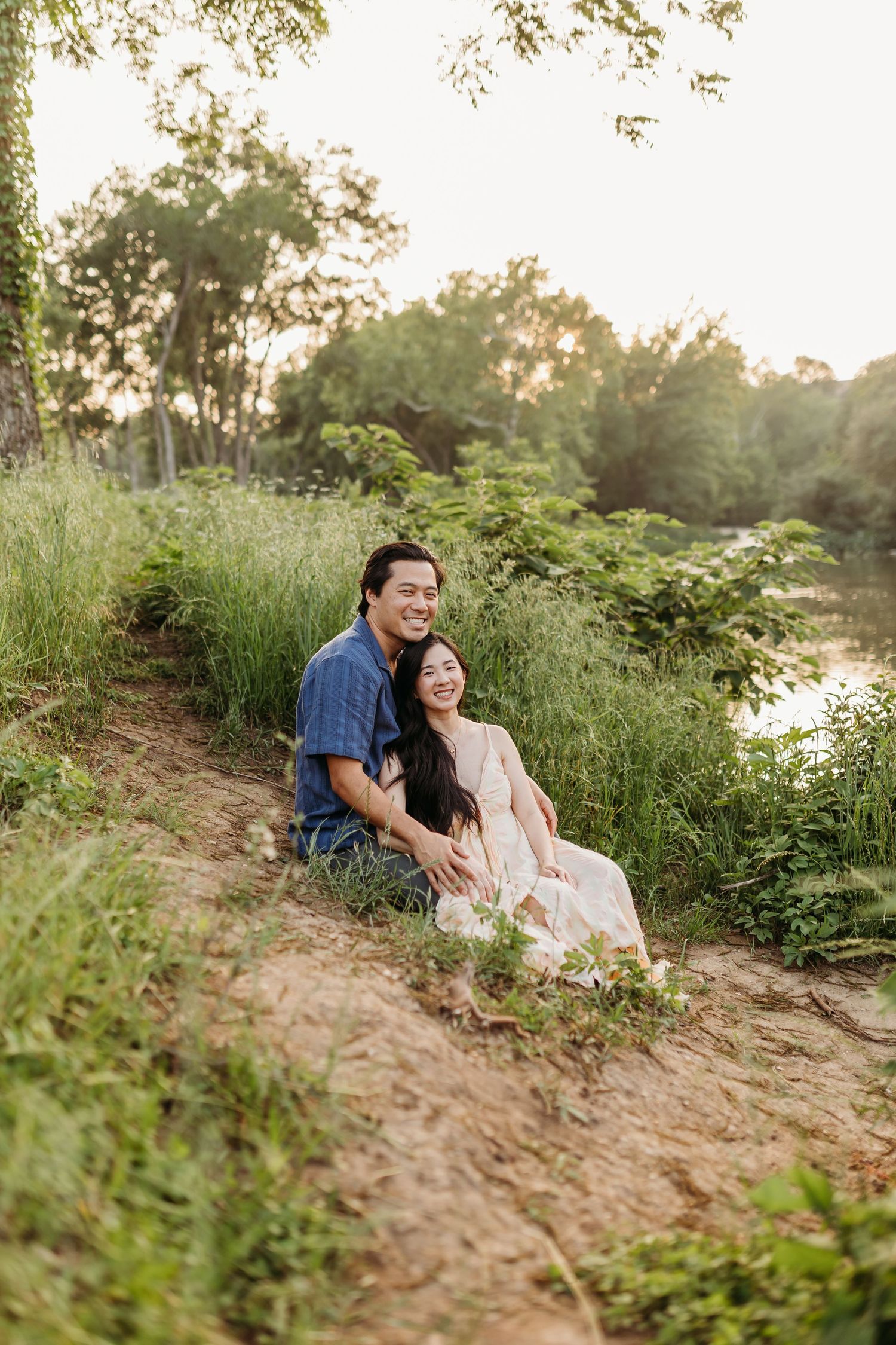 A happy couple sits on a sunlit hillside near a river, surrounded by tall green grass and trees.