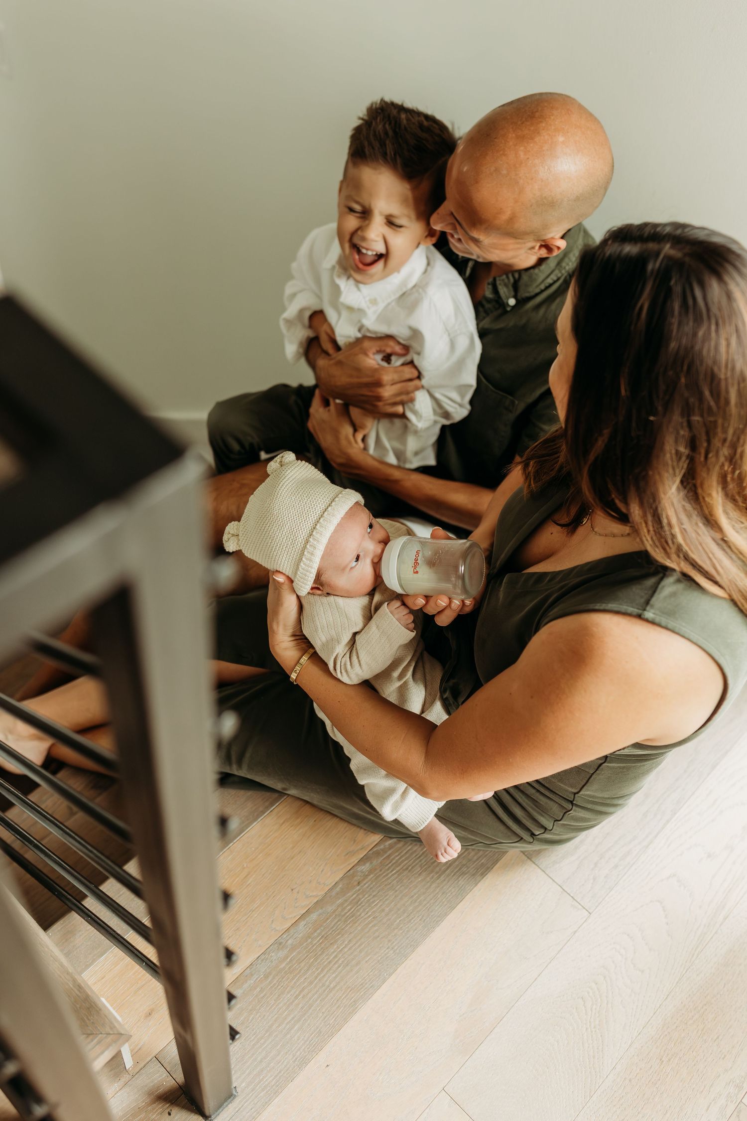 Family sitting on stairs with parents holding two young children, laughing and bottle feeding newborn.