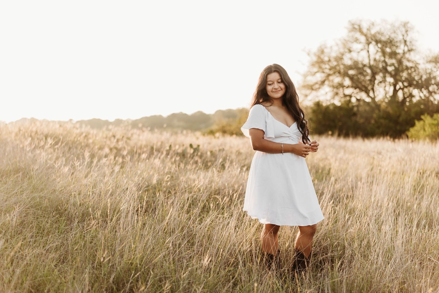 Cheerful young woman in a white dress and boots standing in a golden field during a sunset portrait.