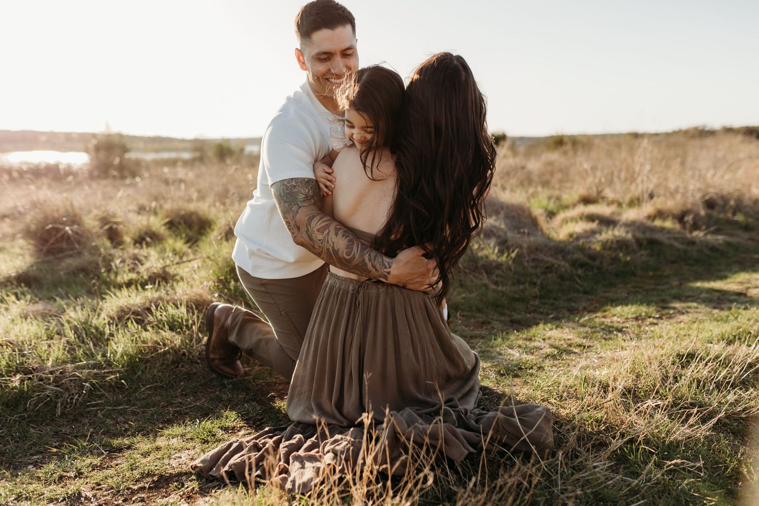 Tattooed man kneeling to embrace a woman in a brown skirt in a sunny outdoor field.