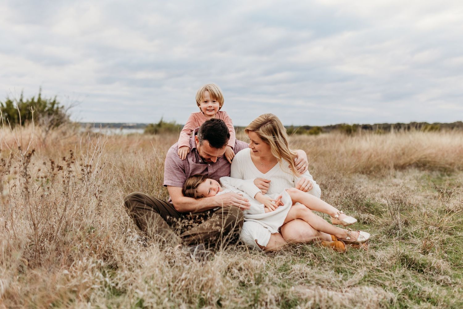 Happy family of four sitting together in golden field with cloudy sky backdrop.