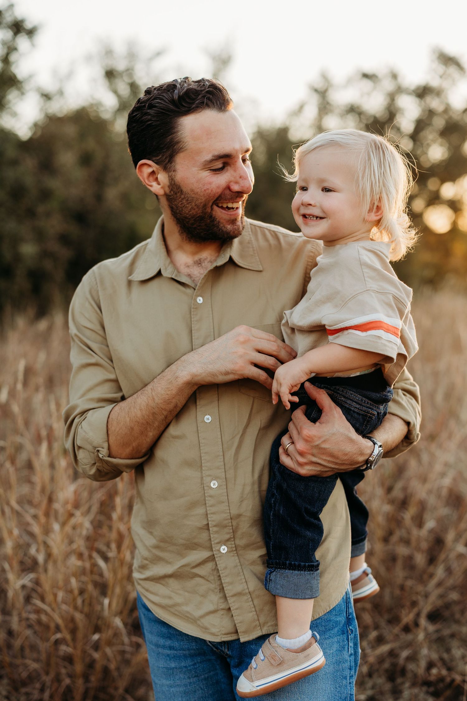 Smiling bearded father holding his laughing toddler in a sun-drenched golden field.