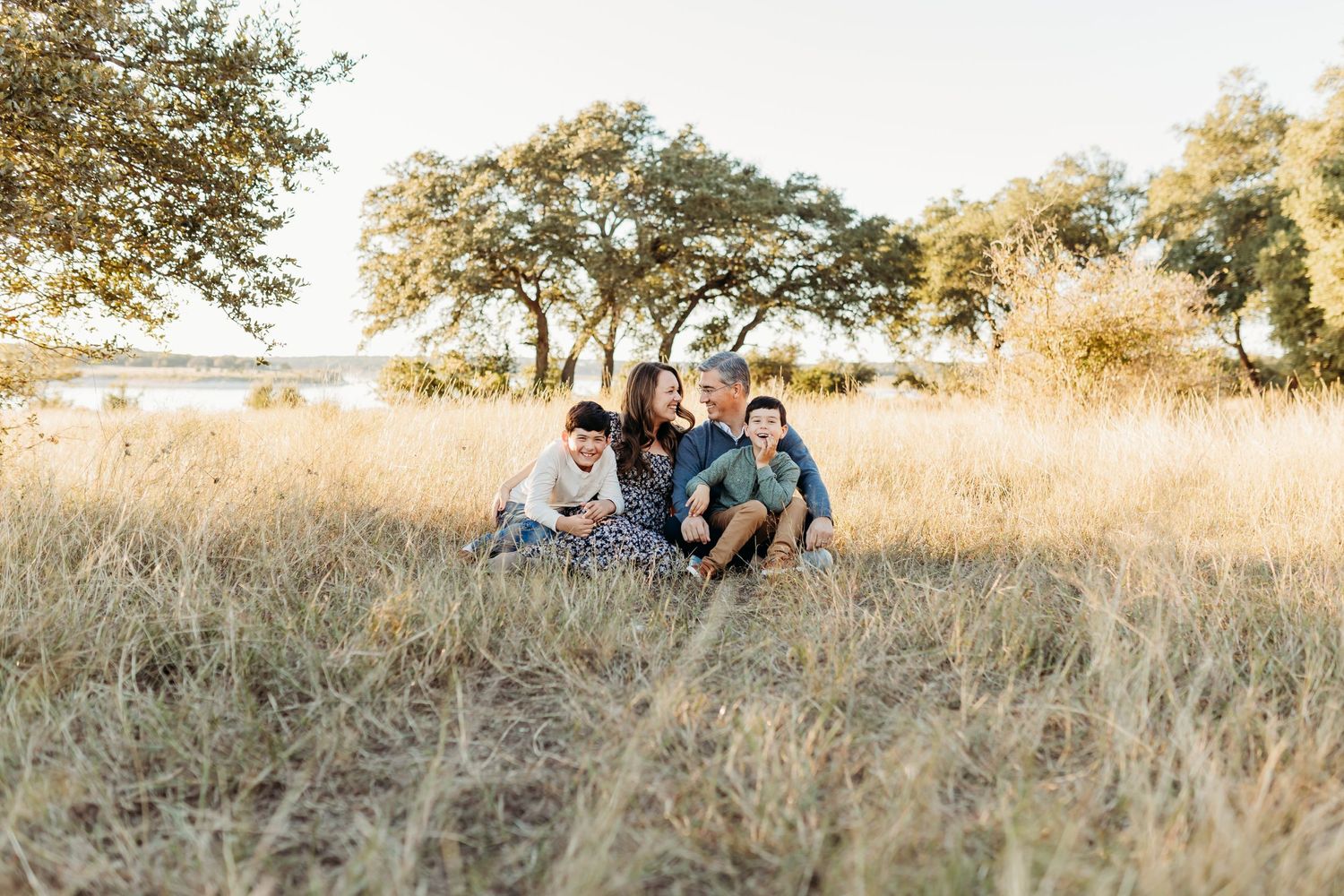 A happy family of five sits together in a sunlit golden grass field with trees in the background.