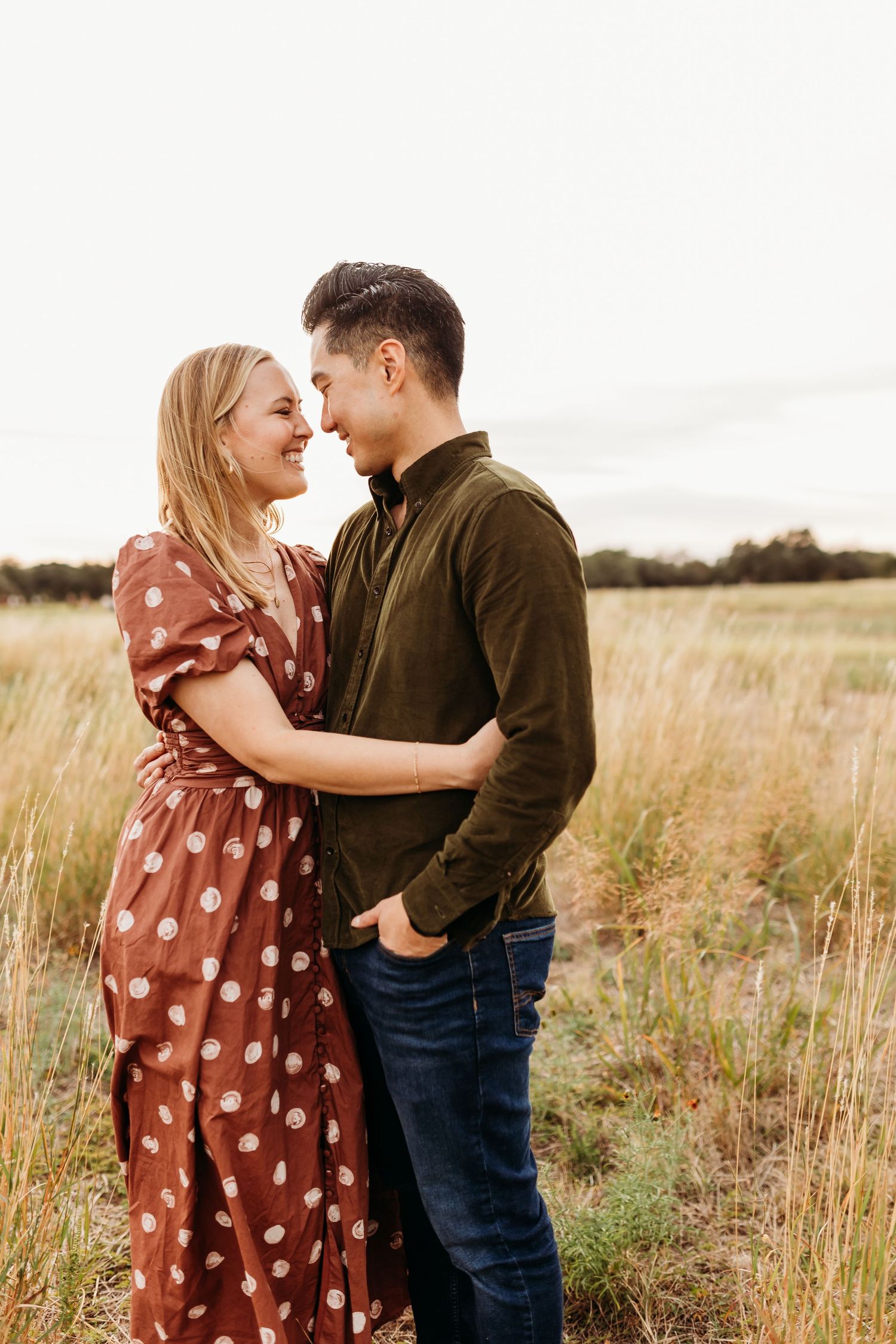 A couple in coordinating earth-toned outfits smile and embrace closely in a golden grassy field at dusk.