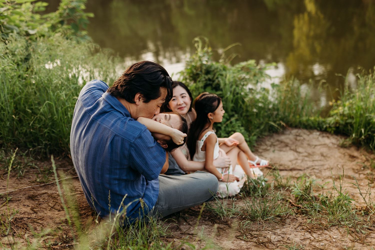 Family of three sitting peacefully by a calm pond surrounded by green grass and nature.