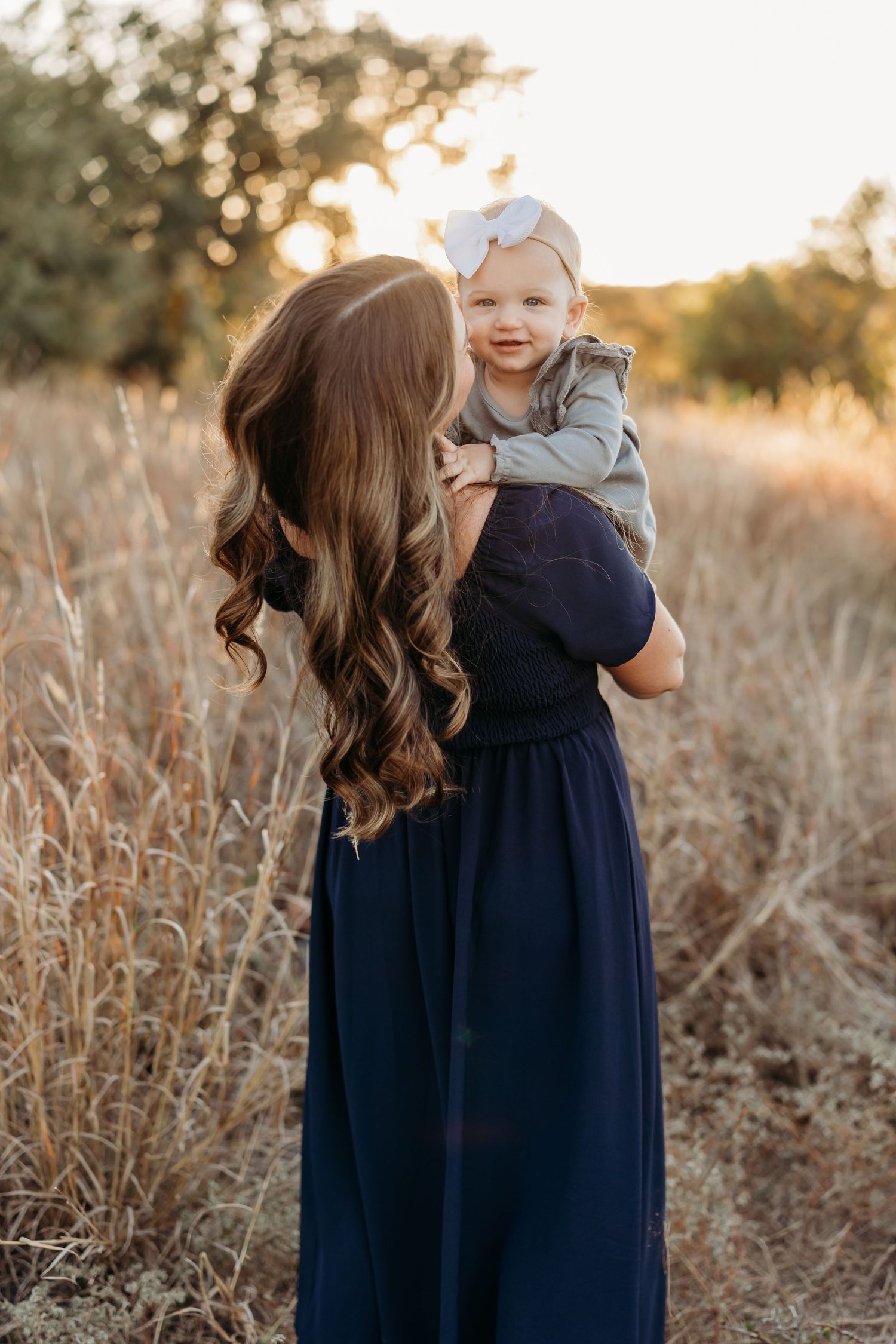 Mother in a navy dress holding her smiling baby girl in a sunlit golden field.