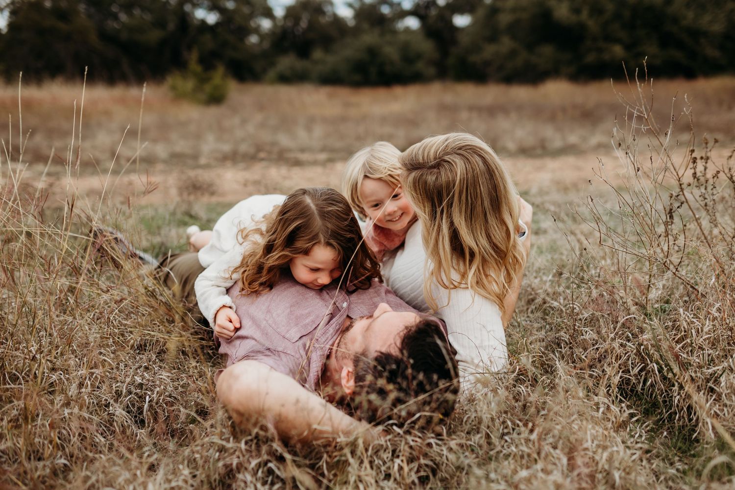 A joyful family laughs together lying in tall dry grass during a relaxed outdoor lifestyle photo session.