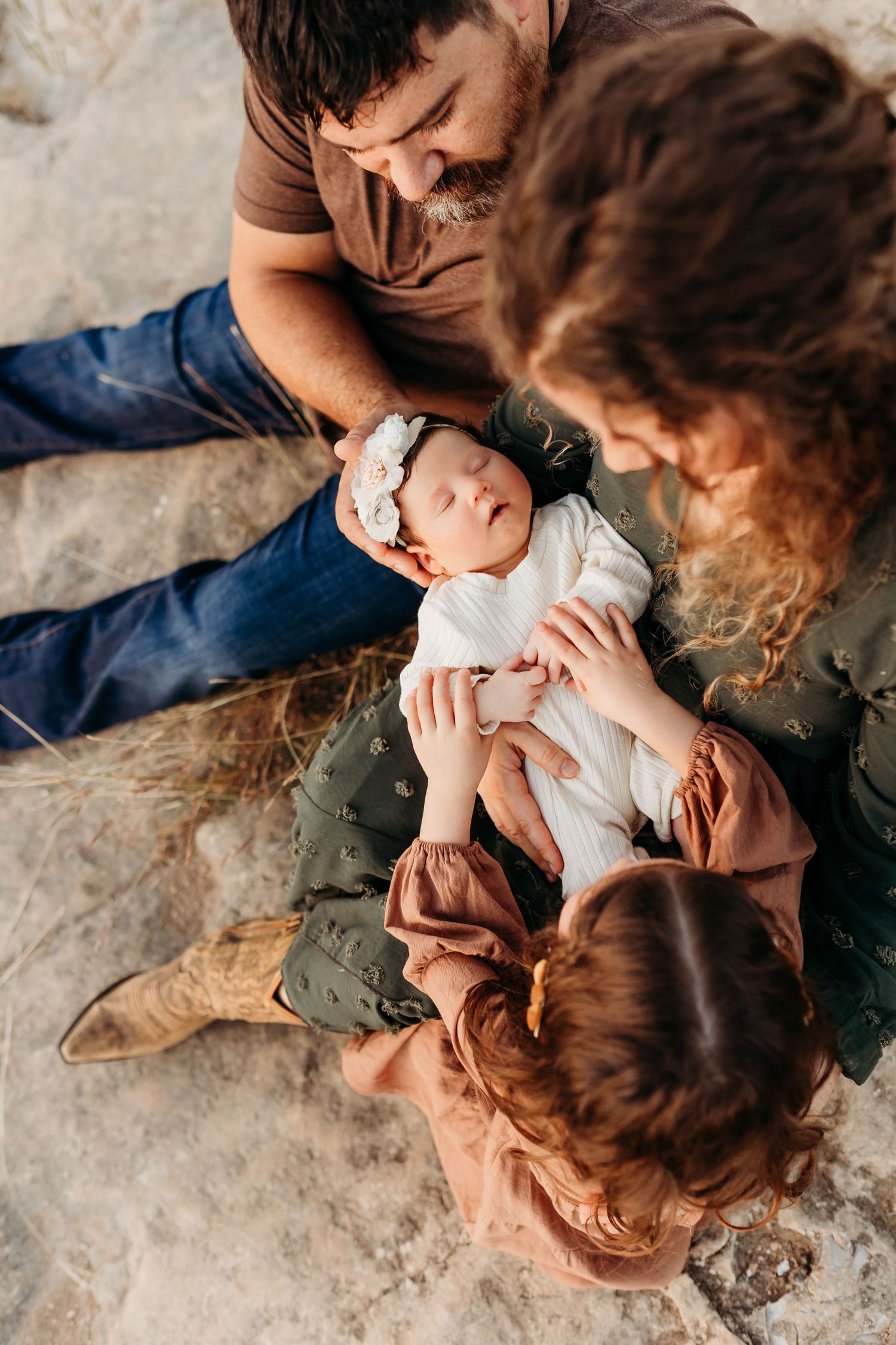 Parents and young daughter admiring sleeping newborn baby wrapped in white on rocky ground outdoors.