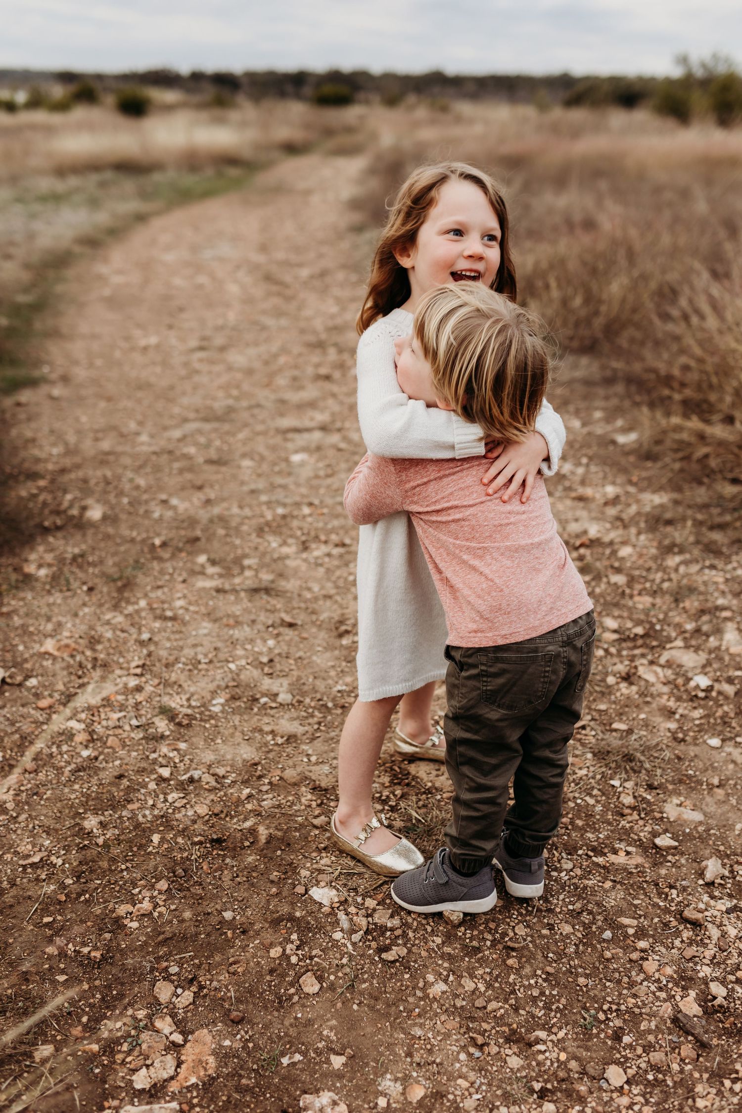 Young girl smiling while hugging little boy on a dirt path in open field.