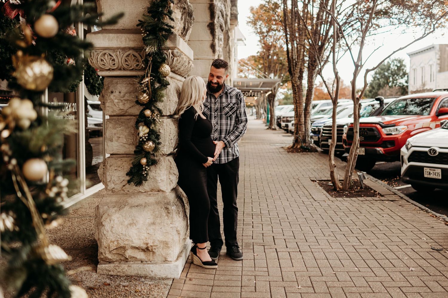 A couple shares a tender moment on a decorated holiday sidewalk beside a festive Christmas tree display.